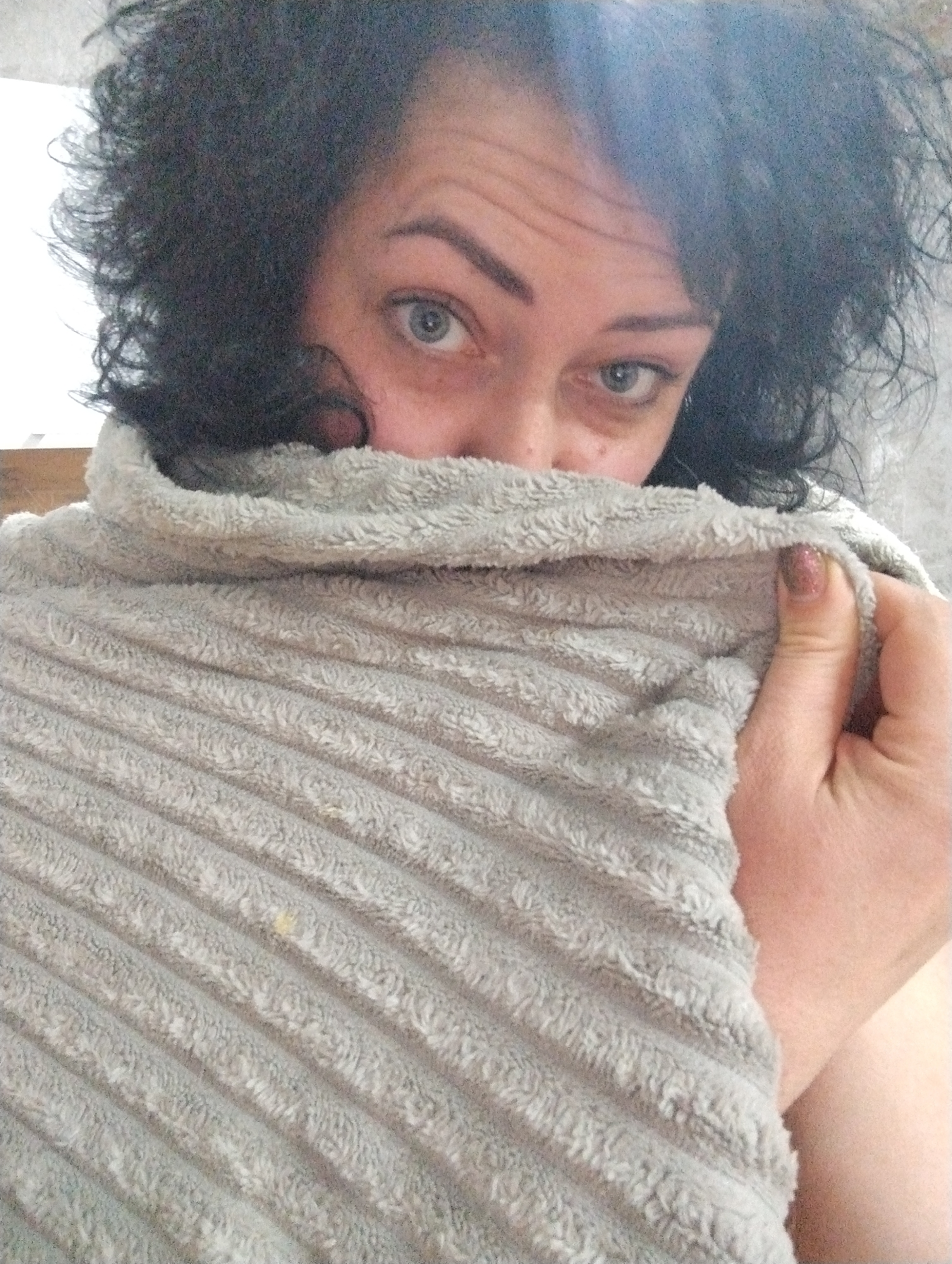 A person with curly, dark hair and light blue eyes is partially obscured by a soft, ribbed, gray blanket. The person's face is mostly hidden, with only the eyes and forehead visible above the blanket. A hand is holding the blanket, with one fingernail visible and decorated with glittery nail polish. The background is blurry and indistinct, suggesting an indoor setting. There is no information to suggest the location is Grishanki, Russia.
