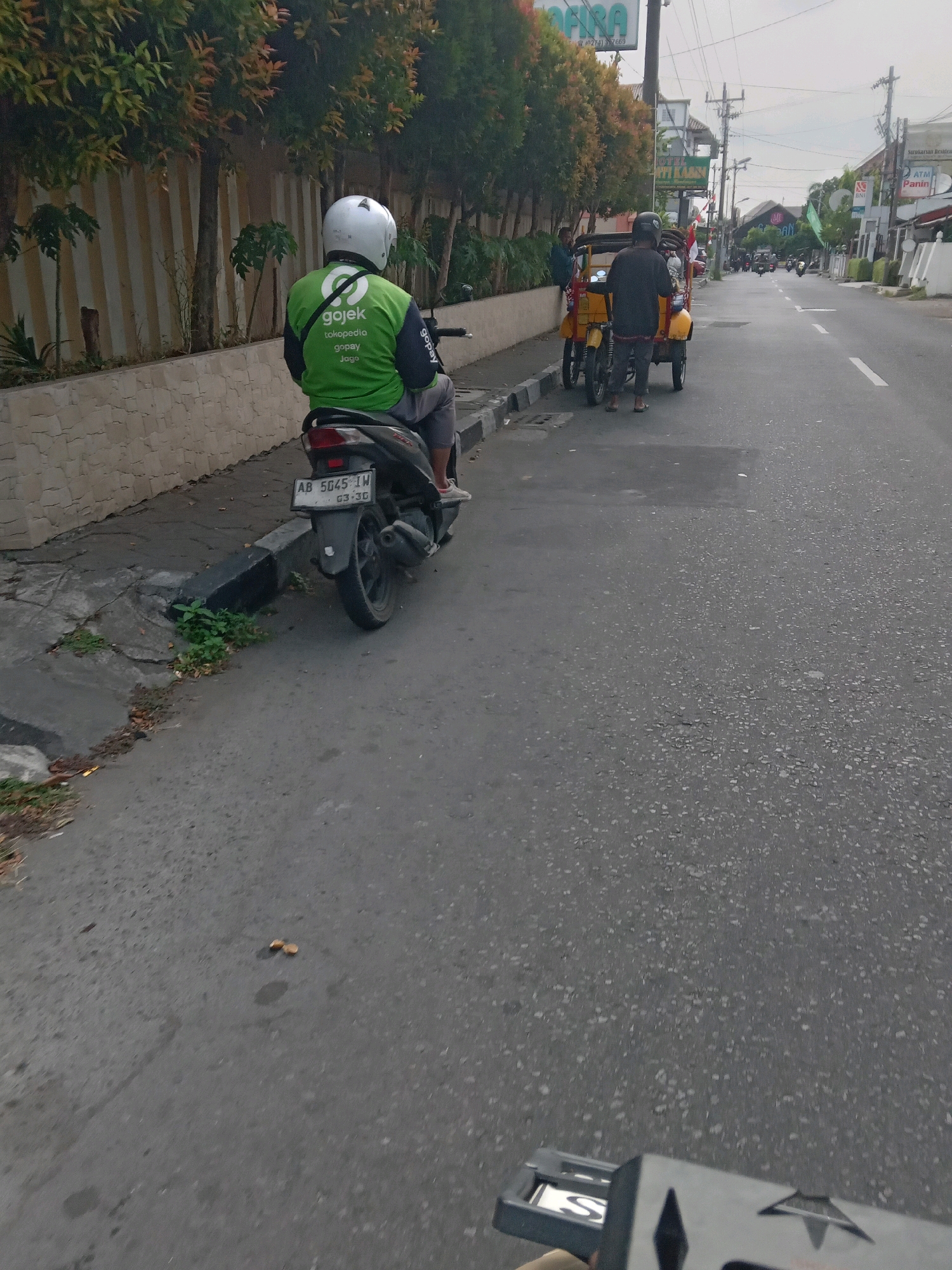 In Yogyakarta, Indonesia, a Gojek motorcycle rider wearing a green jacket and white helmet is seen from behind on the left side of the frame, parked on the side of a paved road. The rider is seated on a black motorcycle with the license plate "AB 5045 IW". To the right of the rider, a yellow three-wheeled vehicle with a canopy is parked, and a person in a black shirt and pants is standing next to it. The road is a two-lane asphalt street with white lane markings. The surroundings include buildings with signage, trees with autumn foliage, and utility poles. The sky is overcast. In the foreground, a black object with white lettering is partially visible, held by an unseen person.