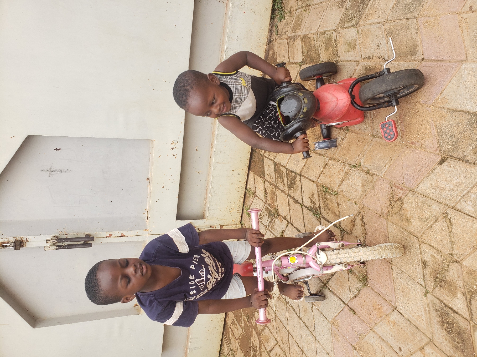 Two young Black children are on tricycles on a paved courtyard in Sabon Tasha, Nigeria. On the left, a child in a dark blue t-shirt with white stripes on the sleeves and light blue shorts is on a pink tricycle with training wheels. The child has their eyes closed and is looking down. On the right, a younger child wearing a black, white, and yellow patterned sleeveless shirt and black patterned shorts is on a red tricycle with training wheels. This child is looking to the right with a smile. The ground is paved with light brown and pinkish square tiles. In the background, a large, light-colored, corrugated metal gate with a padlock is visible. The lighting suggests it is daytime.