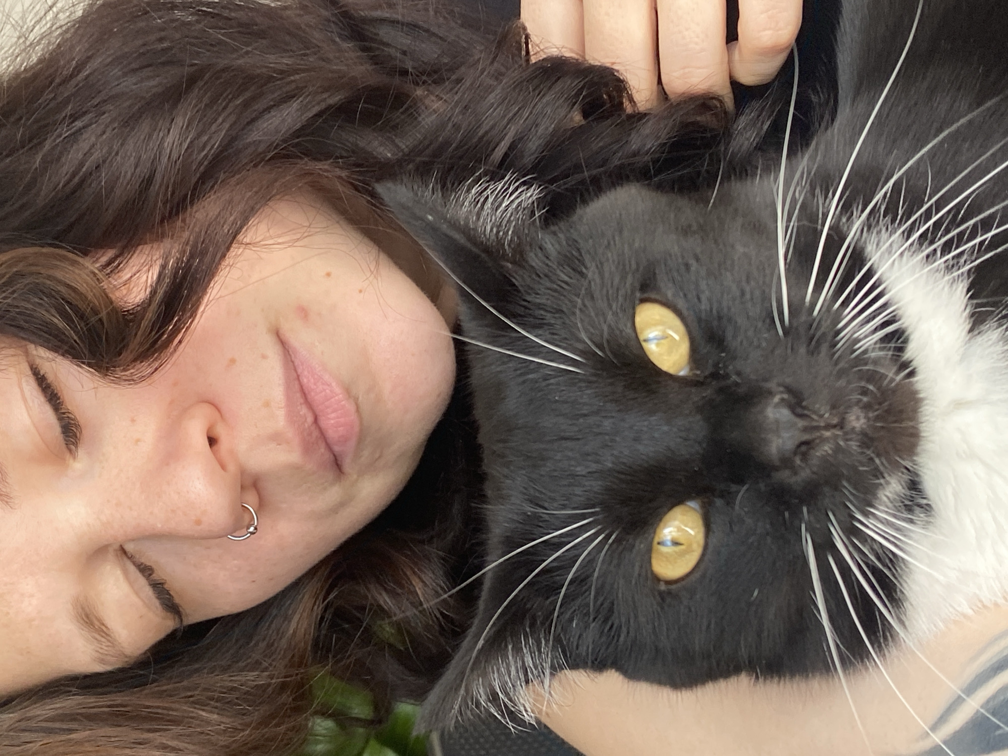 A close-up, selfie-style photograph shows a woman and a black-and-white cat. The woman is fair-skinned with dark, wavy hair and a silver nose ring. Her eyes are closed and her mouth is slightly open. The cat, positioned next to her face, has yellow eyes and long white whiskers. Part of the woman's hand and some dark fabric are also visible in the frame. The location is not discernible from the image.