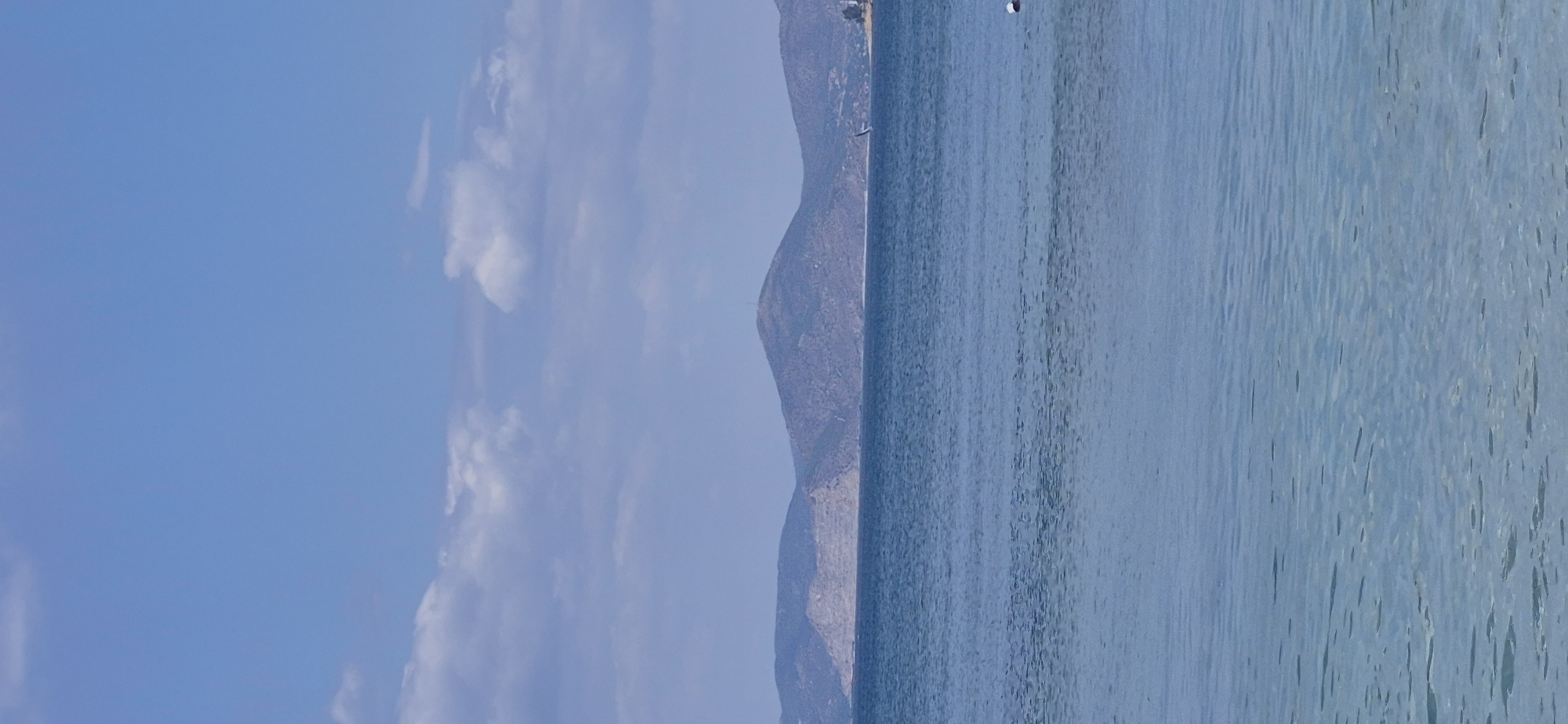 The image depicts a serene seascape with a clear blue sky dotted with white clouds. In the distance, a range of barren, brown hills forms the horizon. The sea in the foreground is calm and transparent, revealing the sandy seabed beneath. There is a small sailboat visible on the water. A person wearing a white cap is partially visible in the water on the right side of the frame, seemingly engaged in swimming or water activity. The scene captures a peaceful moment at a beach, likely in Paleo Faliro, Greece, given the characteristic landscape of the Attica coastline.