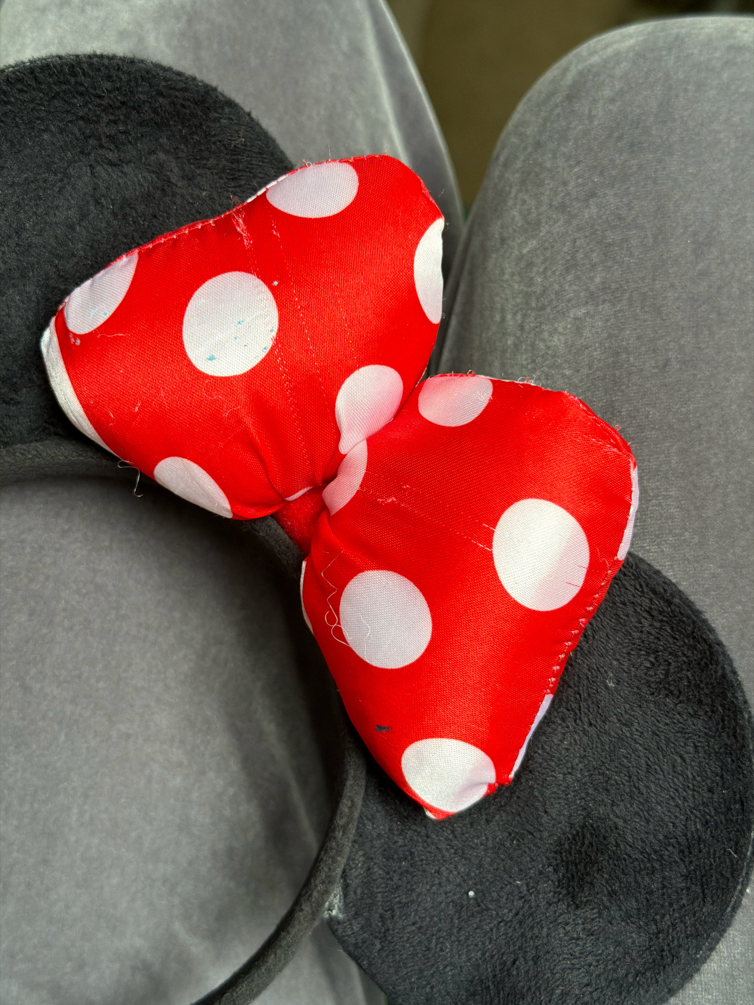 This is a close-up, overhead shot of a pair of Minnie Mouse ears. The ears are black and fuzzy, and the bow is red with white polka dots. The ears are resting on a person's legs, which are covered in gray sweatpants. The lighting is diffused and the background is out of focus, with the subject being the main focus of the image. There are no visible people or animals in the image. The location of Бабініцкі сельскі Савет, Belarus, is not discernible from the image.