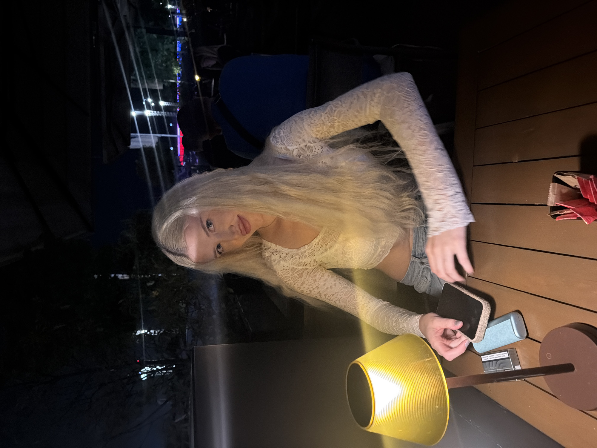 A young woman with long blonde hair sits at an outdoor table, potentially in Belgrade, Serbia. She is wearing a light-colored, long-sleeved lace top and denim shorts. She is holding a black phone in her hands. On the table are a small lamp with a yellow shade, a pack of cigarettes, and a blue case. Other people are visible in the background, and city lights suggest an urban environment. The table has a wooden-planked surface. A box, partially visible, sits near the edge of the table.