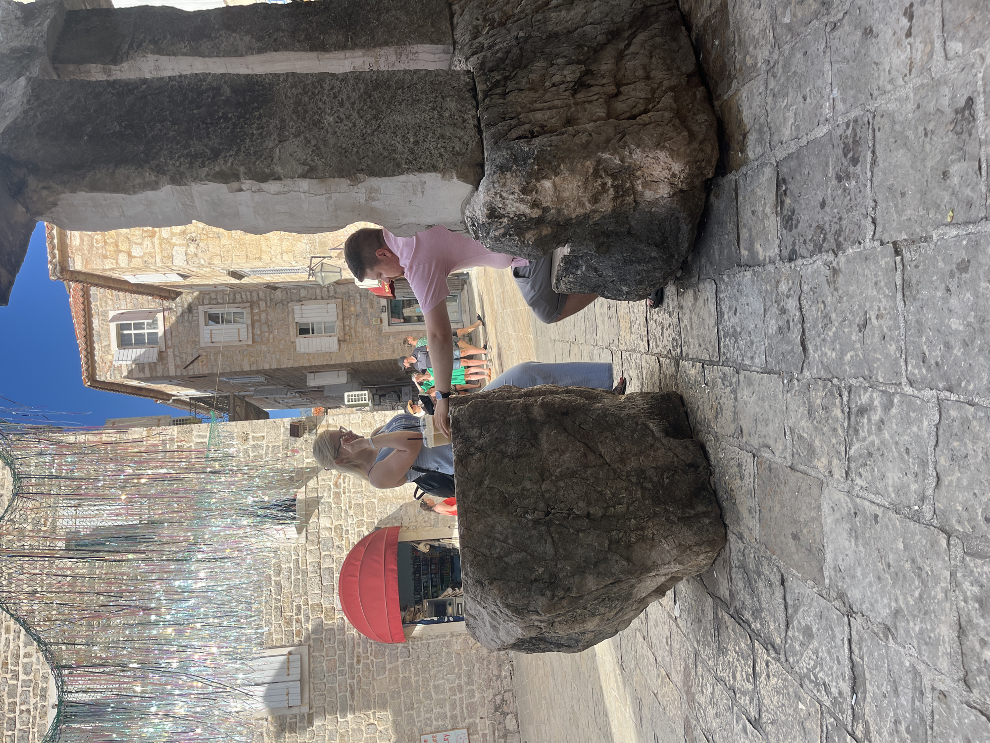 In Budva, Montenegro, two individuals are seated on large boulders, one on either side of what appears to be a stone archway. The woman, with blonde hair and wearing a light blue jumpsuit, is seated on the left boulder and holds a drink with a straw. She is looking down and appears to be touching her face or glasses. The man, on the right boulder and wearing a pink short-sleeved shirt and grey shorts, is leaning back and extending his arm. Behind them, a narrow street is visible with stone buildings featuring red-tiled roofs and shutters. To the left, a small shop with a red awning is open. Overhead, decorative, iridescent streamers hang from a building. The ground is paved with large, grey cobblestones, and some of the stones appear wet. The overall scene suggests a sunny day in a historic town.
