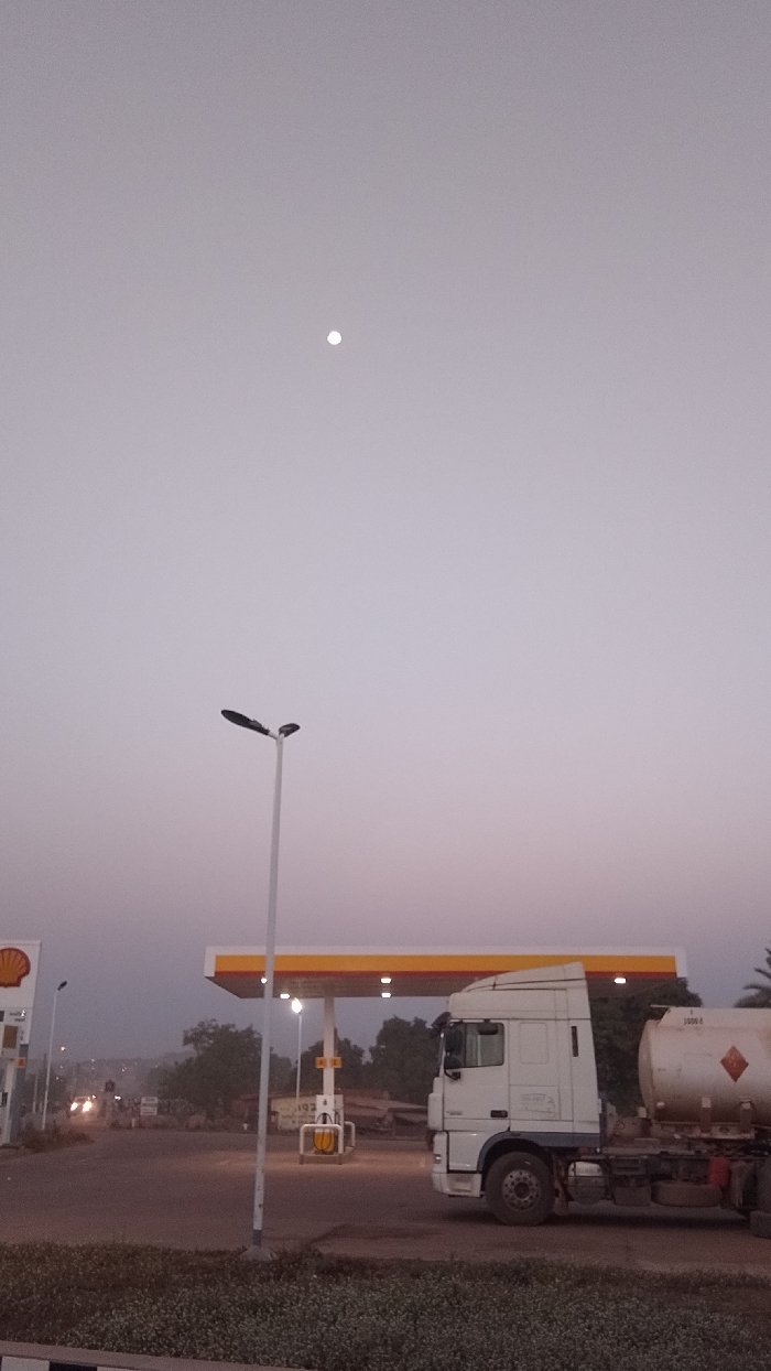 A fuel truck sits parked at a Shell gas station under a pale sky with a bright moon. The canopy of the station is brightly lit. A tall street lamp stands nearby. Across the road, blurry lights suggest other vehicles. The scene appears to be at dusk or dawn, characterized by muted colors and a quiet atmosphere.