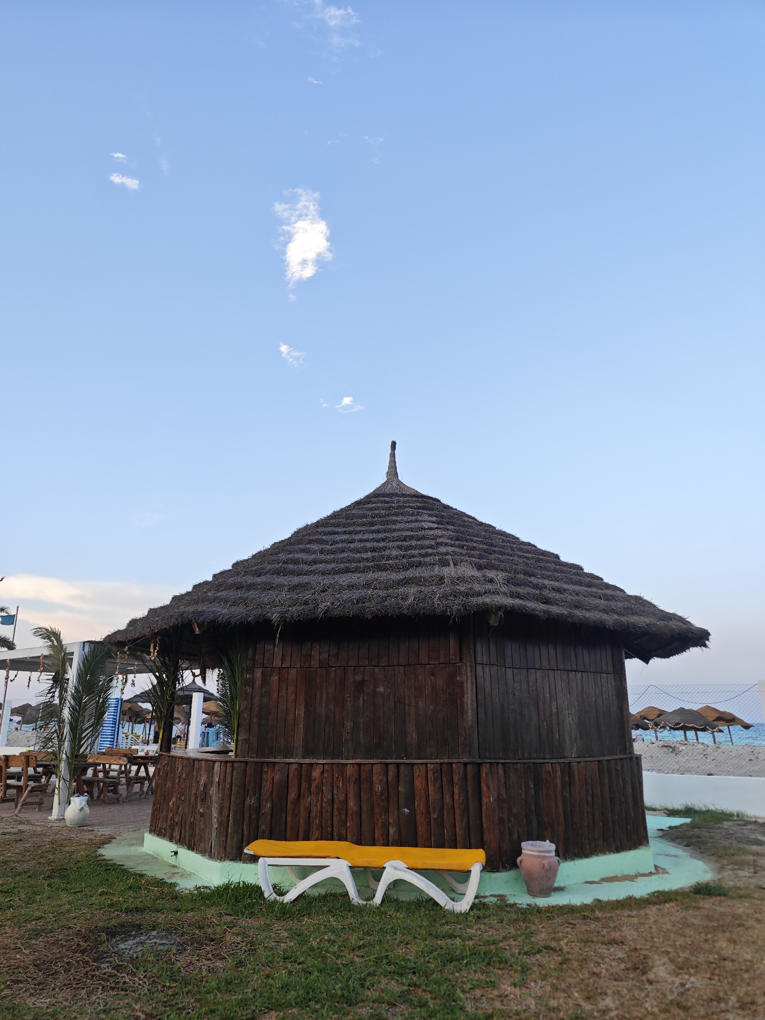 This is a photograph taken in Mahdia, Tunisia. The image captures a beachfront scene with a focus on a round, wooden structure topped with a thatched roof. In front of the structure, there is a yellow lounge chair and a terracotta pot. The background reveals the beach with a fence, more thatched umbrellas, and the blue sea under a pale blue sky with sparse clouds. To the left of the wooden structure, there are tables and chairs, suggesting an outdoor seating area, possibly for a restaurant or cafe. Palm trees are also visible, adding to the tropical ambiance. The overall impression is one of a relaxed, coastal environment.