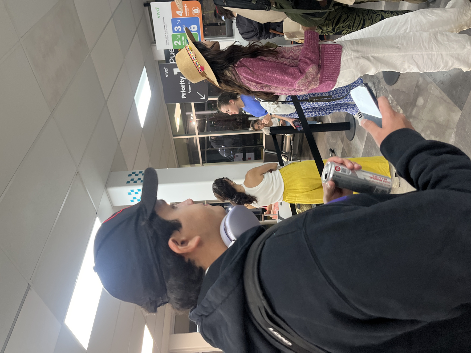 Here is a description of the image:
The image shows a slightly rotated, close-up view of a crowded airport terminal. In the foreground, a person wearing a black baseball cap, black hoodie, and gray headphones is holding a can of soda and a smartphone, seemingly taking a picture. The foreground subject is predominantly out of focus.
Further into the frame, several people are standing in a line, presumably waiting. One person wears a pink sweater, white pants, and a straw hat. Behind them, other people are wearing blue shirts and dresses.
Signs are visible in the background with the words "Priority" and "Viva," along with colored icons representing numbers 2, 3, and 4, with labels "verde," "azul," and "naranja" beneath them. Based on the signs in the image, the location is in the city of Puerto Escondido, Mexico.