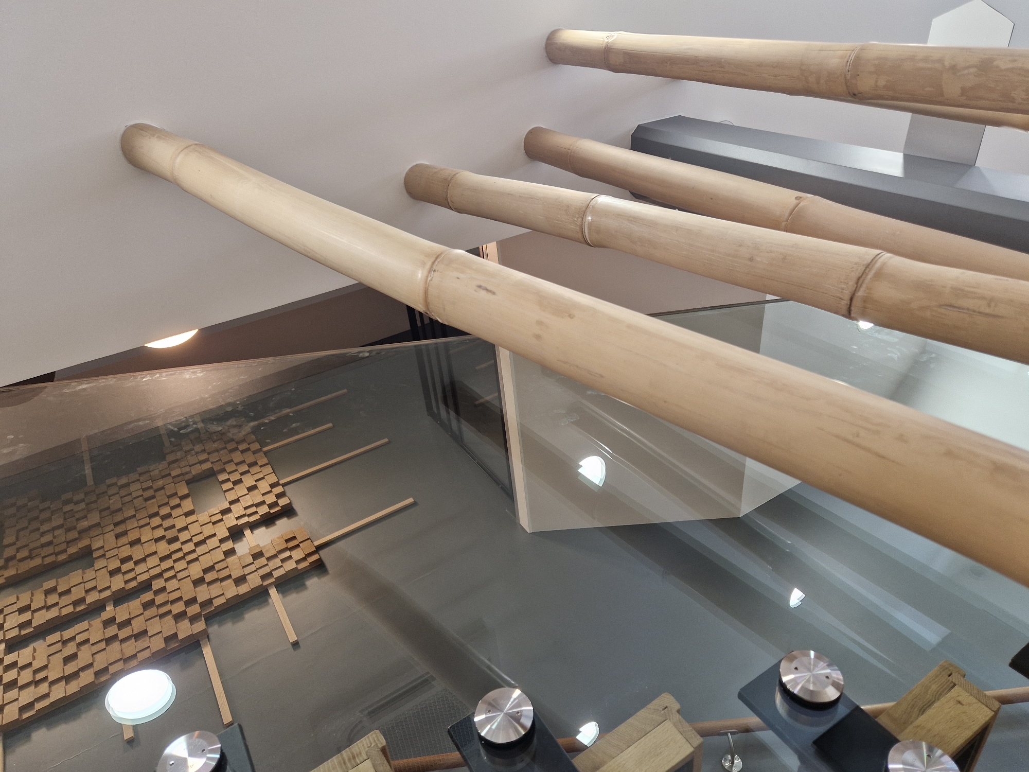 The image depicts an interior space with a modern aesthetic, featuring elements of natural materials and reflective surfaces. Prominently visible are several large, light-colored bamboo poles, angled upwards towards a white ceiling. Some of these poles appear to be structural supports, with visible nodes characteristic of bamboo.
On the left side of the image, a wall is covered with a textured, wood-like material arranged in a grid of small cubical blocks. A circular light fixture with a white diffuser is embedded in this wall. Adjacent to this wall is a large, highly reflective surface, likely glass, which mirrors parts of the bamboo poles and the ceiling, creating a sense of depth and spatial complexity.
Several circular, metallic fixtures with a brushed finish are attached to the reflective surface, appearing to hold or support other elements. Below these fixtures, wooden components that resemble shelves or display units are partially visible.
In the background, a dark grey angular structure is visible, possibly a pillar or part of the building's architecture. The overall lighting in the scene is soft, with some sources contributing to the reflections on the glass. The image does not contain any people.
This scene is located in the **Médiathèque La Canopée, Antony, France.**