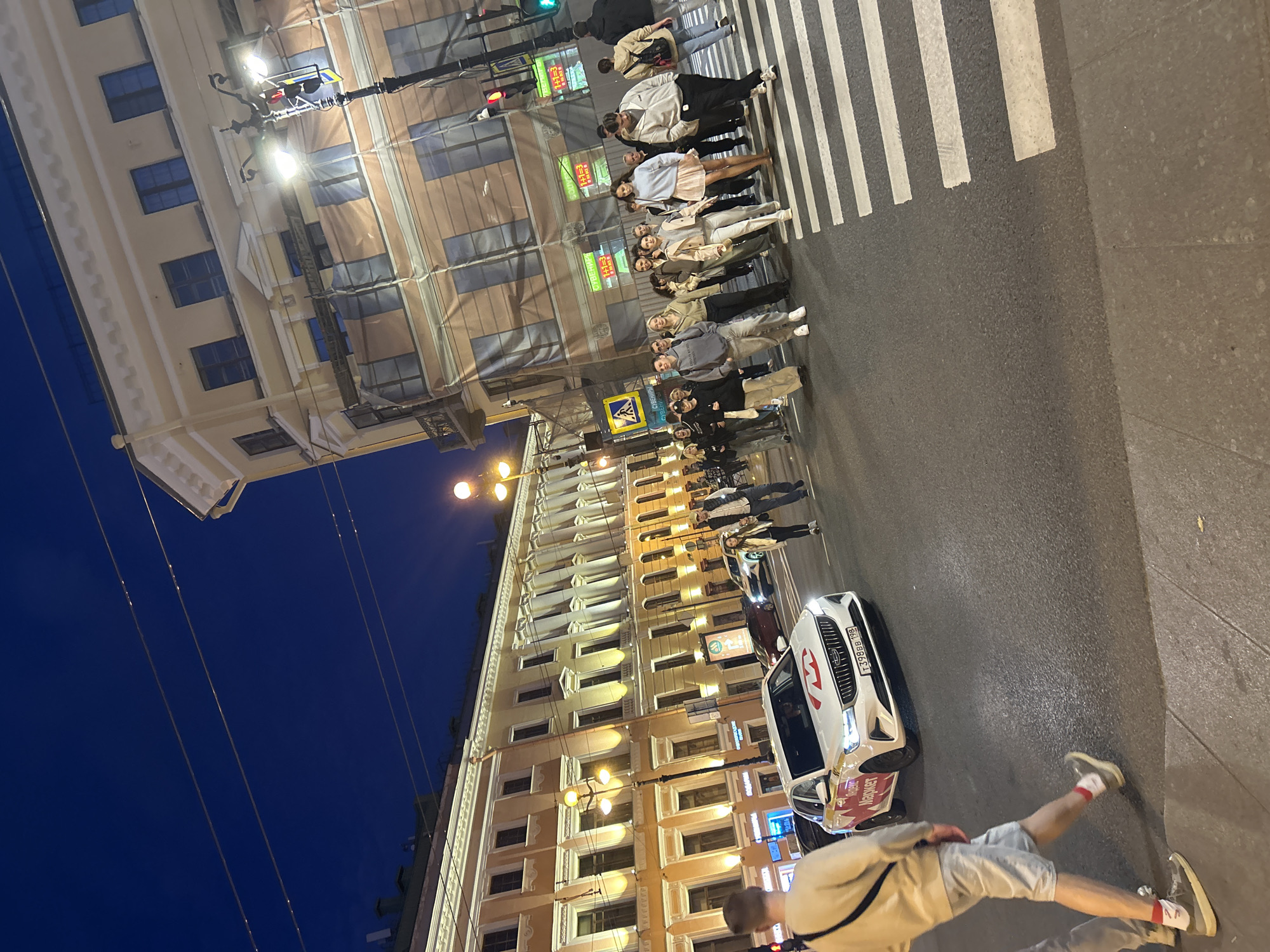In an urban street scene at dusk, a white sedan with red and yellow branding is stopped at a crosswalk. The car displays a license plate reading "T398BB 198". A group of pedestrians, predominantly young adults dressed in casual attire, are crossing the street. They are illuminated by streetlights and the warm glow emanating from the windows of adjacent buildings. The buildings, featuring classical architectural elements, are lit up, contrasting with the darkening blue sky. Traffic lights are visible, and overhead wires crisscross the sky. The ground is paved with asphalt, and the crosswalk is clearly marked with white stripes. The overall atmosphere suggests a busy city street in the evening.