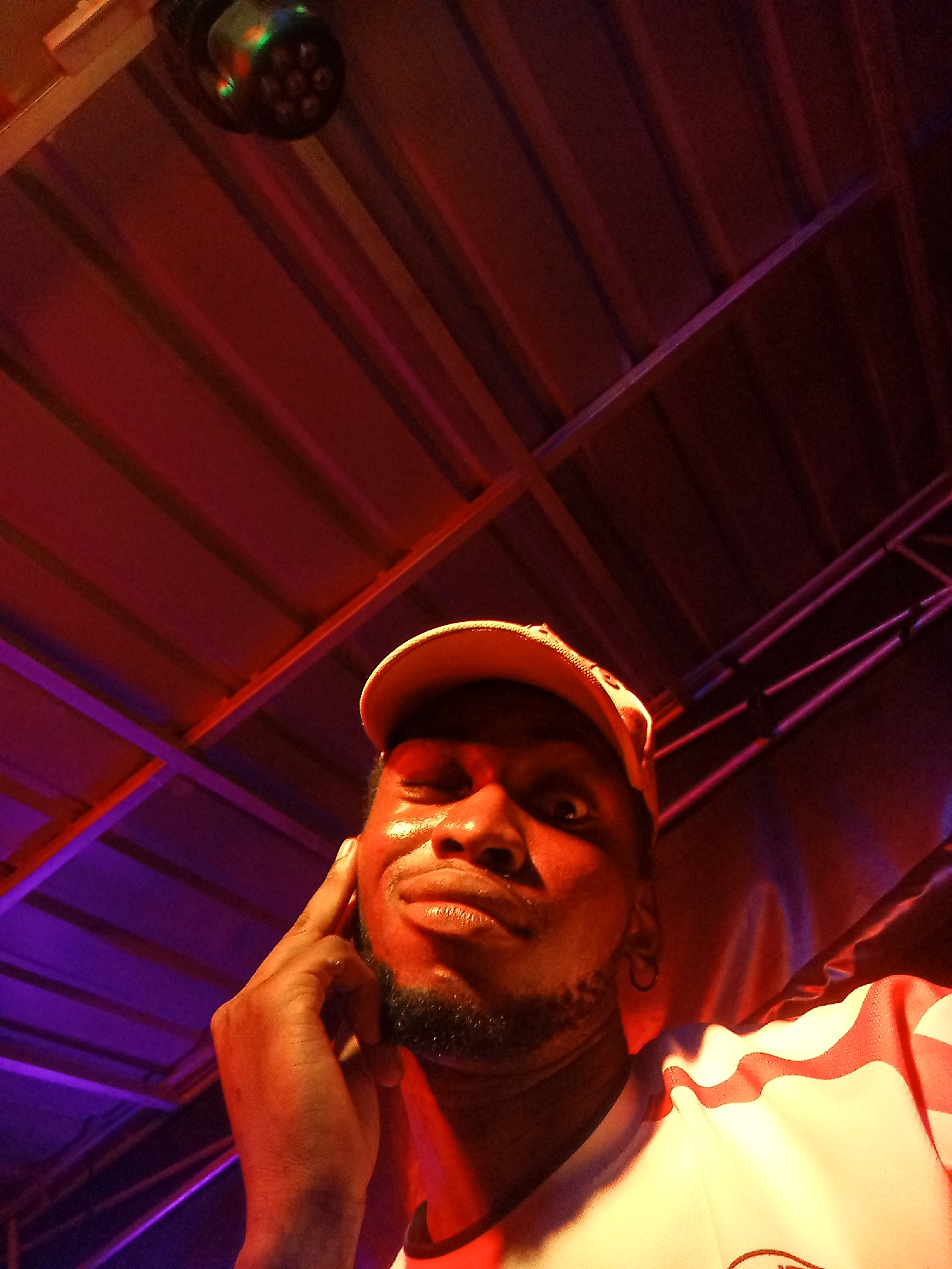 A man is posing for a selfie in a room with red and purple lighting. The ceiling is visible in the background, with metal beams and a multi-bulb light fixture. The man is wearing a cap and a white shirt with red stripes. He has a beard and is holding his hand up to his face, seemingly winking. The location cannot be determined.
