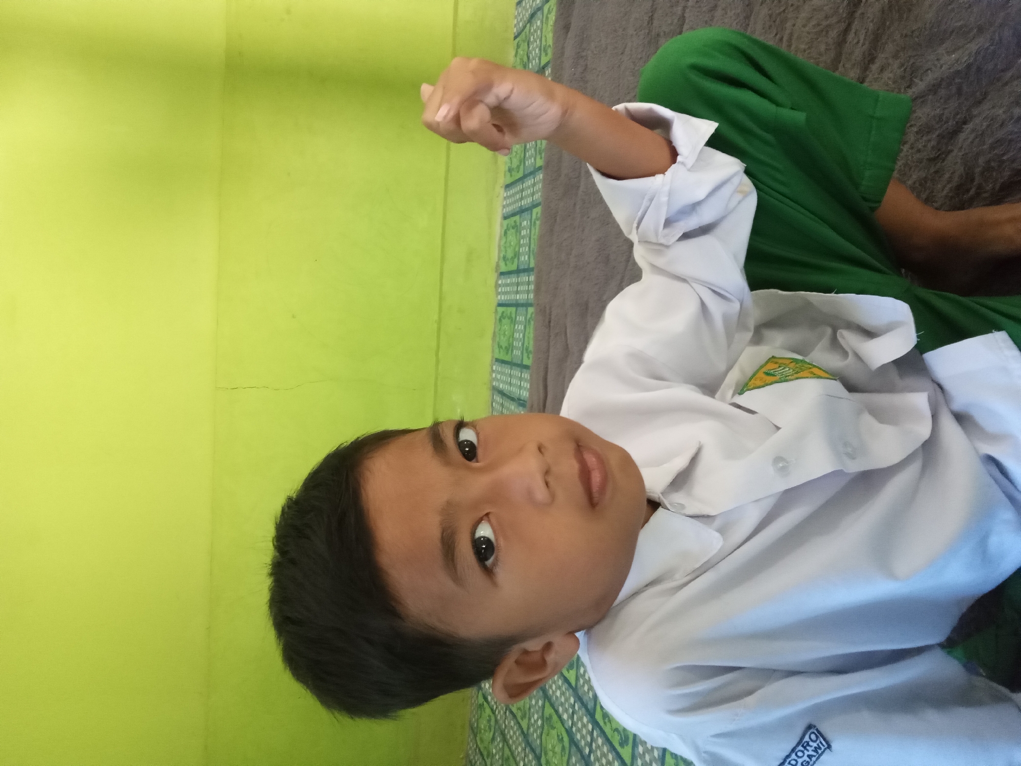 A young boy in a white collared shirt and green pants sits on a mat in front of a lime green wall. The boy has dark hair and brown eyes and is looking at the camera. His left hand is raised in a fist. The mat has a pattern of green squares. The wall has a slight texture and a visible horizontal crack. The scene appears to be indoors, possibly in a bedroom or classroom. The location is identified as Kedunggudel, Indonesia.