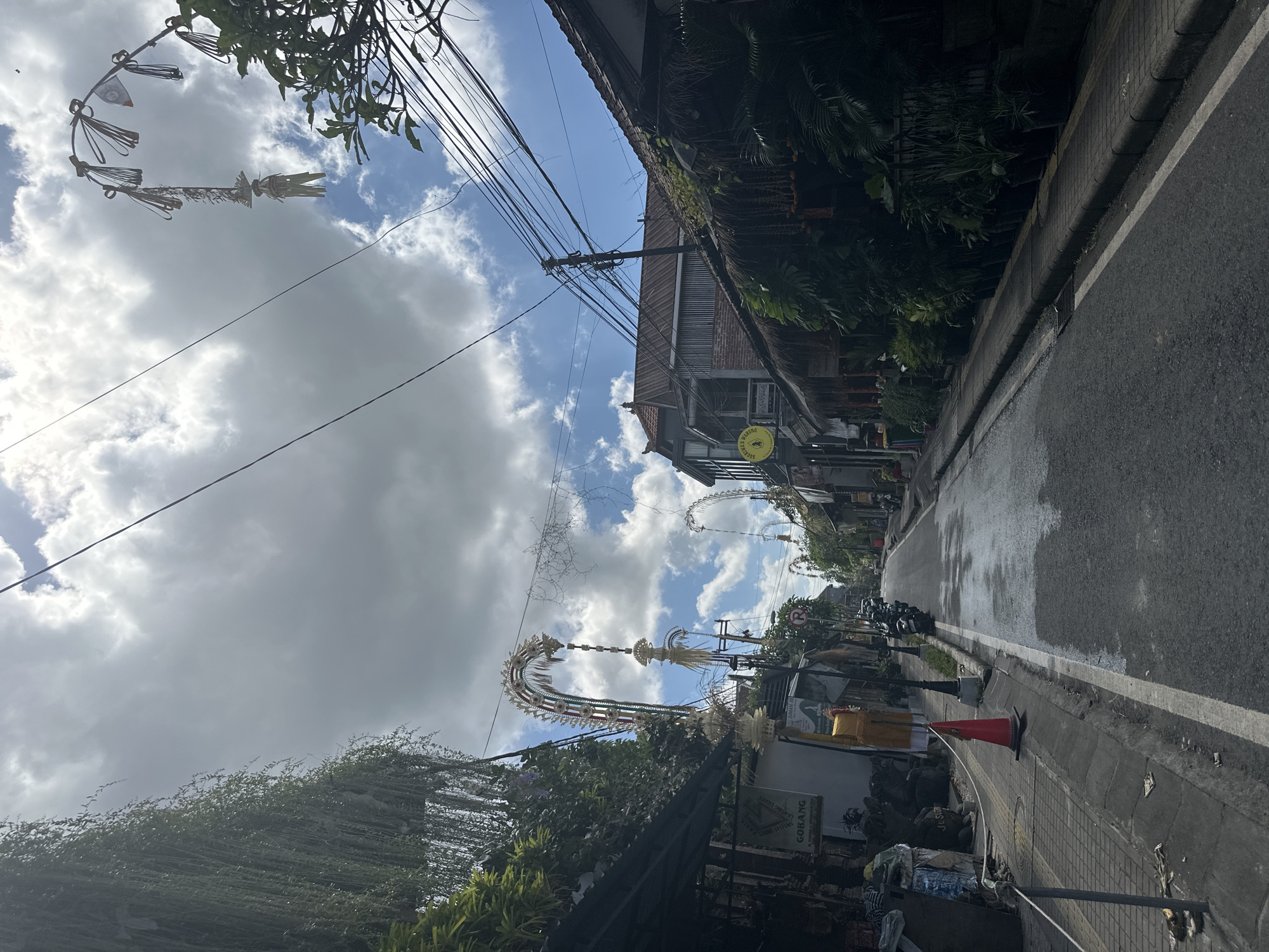 The image depicts a street scene in Ubud District, Indonesia, characterized by overcast skies with patches of blue. The road is paved and appears wet in places, suggesting recent rain or street cleaning. On the left side of the street, there are buildings with awnings and hanging plants, with a sign that reads "GOBANG" visible. A red traffic cone is placed on the sidewalk. Further down the street on the left, a few motorcycles are parked.
On the right side of the street, there are more buildings with traditional Balinese architecture, featuring thatched roofs and lush greenery. Decorative arches, characteristic of Balinese street decorations, are suspended across the road, adorned with tassels and small flags. Overhead, a tangle of power lines crisscrosses the sky. The overall impression is of a vibrant and culturally rich urban environment.