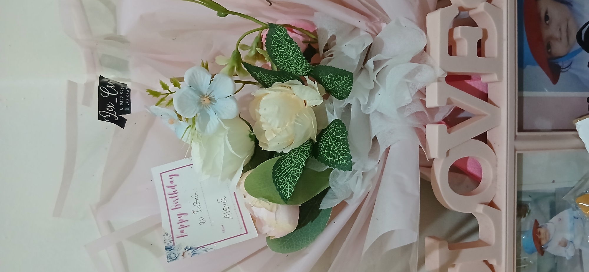 The image shows a bouquet of artificial flowers, predominantly white and cream colored, with accents of light blue and pink. A white card with a floral border is attached to the bouquet. The text on the card reads "Happy Birthday" in pink script, followed by "bu Indah:" and "FROM Alexa" in black script. A black sticker with white text, "lex_craft", and a phone number "0812 536..." and "@lex_craft" is affixed to the packaging of the bouquet, which is made of pink and white tissue paper. Below the bouquet, a pink wooden cutout spelling "LOVE" is visible. Behind the cutout are framed photographs of children. The image appears to be taken indoors, possibly in a home setting. There is no visible information in the image that indicates a specific location in Samarinda, Indonesia.