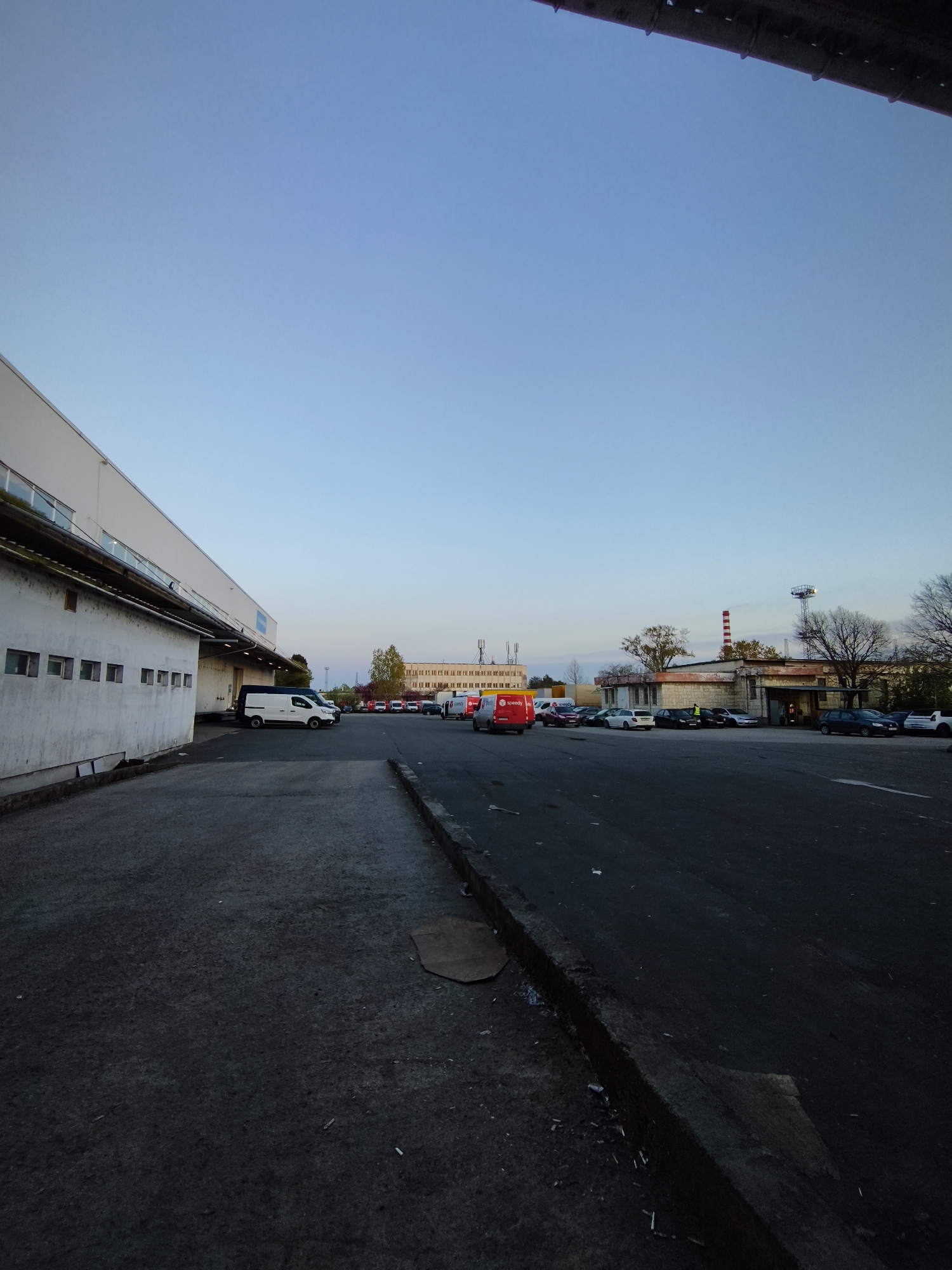This media file depicts an industrial or commercial area in Burgas, Bulgaria, characterized by large buildings and a parking lot, likely during early morning or late afternoon under a clear, pale blue sky. The ground in the foreground consists of asphalt with a concrete curb running along the right side, showing scattered litter and a piece of cardboard.
To the left, a long, white industrial building or warehouse features multiple loading bays and small, rectangular windows. Attached below its roofline is an older, weathered single-story structure. Several white vans are parked near these buildings.
In the middle distance, an array of cars and vans are parked on an asphalt lot. A striking red delivery van, prominently displaying the "Speedy" logo, is visible among them. A person wearing a high-visibility vest stands near these vehicles. Beyond the parking area, an older, single-story building with a flat roof and a light-colored facade is present. Behind this, a distinctive red and white striped chimney stack rises, alongside a communication tower with lights. Further back, a larger multi-story building can be seen. Trees, some deciduous and bare, others still retaining leaves, are interspersed throughout the scene.
The visible "Speedy" logo confirms the presence of a logistics or courier service, which, given the location context, is a Bulgarian company. The overall atmosphere is functional and practical, reflecting an active commercial or industrial environment.