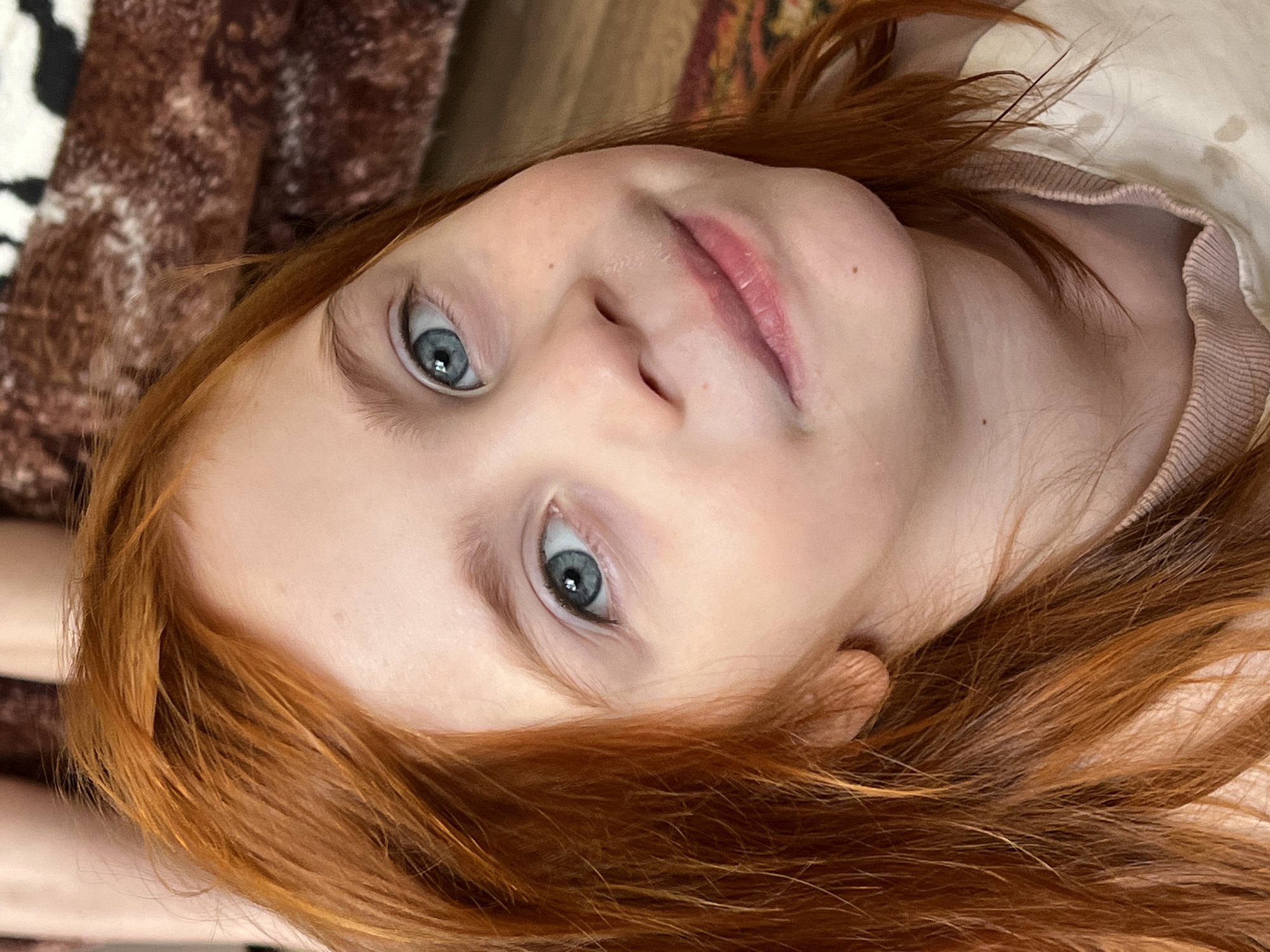 This close-up, vertically oriented image features a young child with bright red hair. The child's face is tilted upwards, with their blue eyes looking directly at the camera. Their lips are slightly parted, and they appear to have a light pink lipstick or gloss on. The child's skin is fair with a few freckles visible on their nose. Their hair is a vibrant auburn, framing their face and falling around their shoulders. They are wearing a light-colored, possibly cream or beige, long-sleeved shirt with a ribbed neckline.
The background is soft and out of focus, but suggests a home environment. To the left, a textured, brown and white patterned blanket or throw is visible. To the upper right, a hint of a patterned rug or fabric with earthy tones can be seen. The lighting appears to be soft, suggesting it might be daytime, possibly indoors with natural light. The overall mood of the image is intimate and personal, capturing a candid moment. There is no visible text in the image. The location context provided is Panteleevka, Russia, which is not directly discernible from the visual elements of the photograph itself.