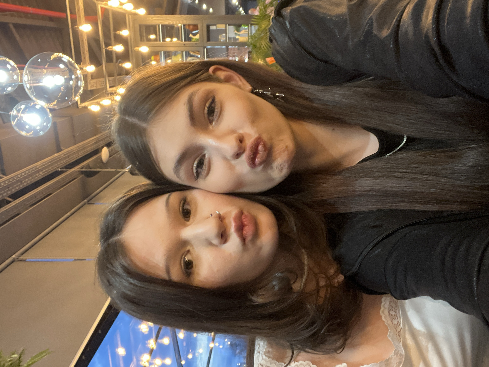 The image is a vertical selfie featuring two young women, taken indoors in what appears to be an evening or nighttime setting. Both women are positioned close together, with one's head resting near the other's, and both are looking towards the camera with puckered lips, making a "kissing face" or pout.
The woman positioned higher and slightly behind has long, dark hair parted in the middle and light-colored eyes. She is wearing a dark top, and a decorative earring, possibly star-shaped, is visible on her right ear.
The woman in the lower, foreground part of the image also has long, dark hair and light-colored eyes. She sports a double nose piercing, consisting of two small studs. She wears a dark top, under which a white garment with a delicate lace trim is visible, possibly a camisole or undershirt.
The background indicates an atmospheric indoor location, potentially a restaurant, bar, or cafe. Notable features in the background include various light fixtures: several large, spherical, reflective lights resembling modern pendant lamps or decorative orbs, and numerous smaller, exposed filament bulbs arranged in frames or strung across the space. These lights emit a warm glow. Metal structural elements, such as beams or frames, are also visible. A blurred area in the lower background suggests a window looking out onto more lights or a reflection.
The overall emotion conveyed is playful and friendly, capturing a casual, intimate moment between the two individuals. The location context provided is Saint Petersburg, Russia. There is no visible text within the image.