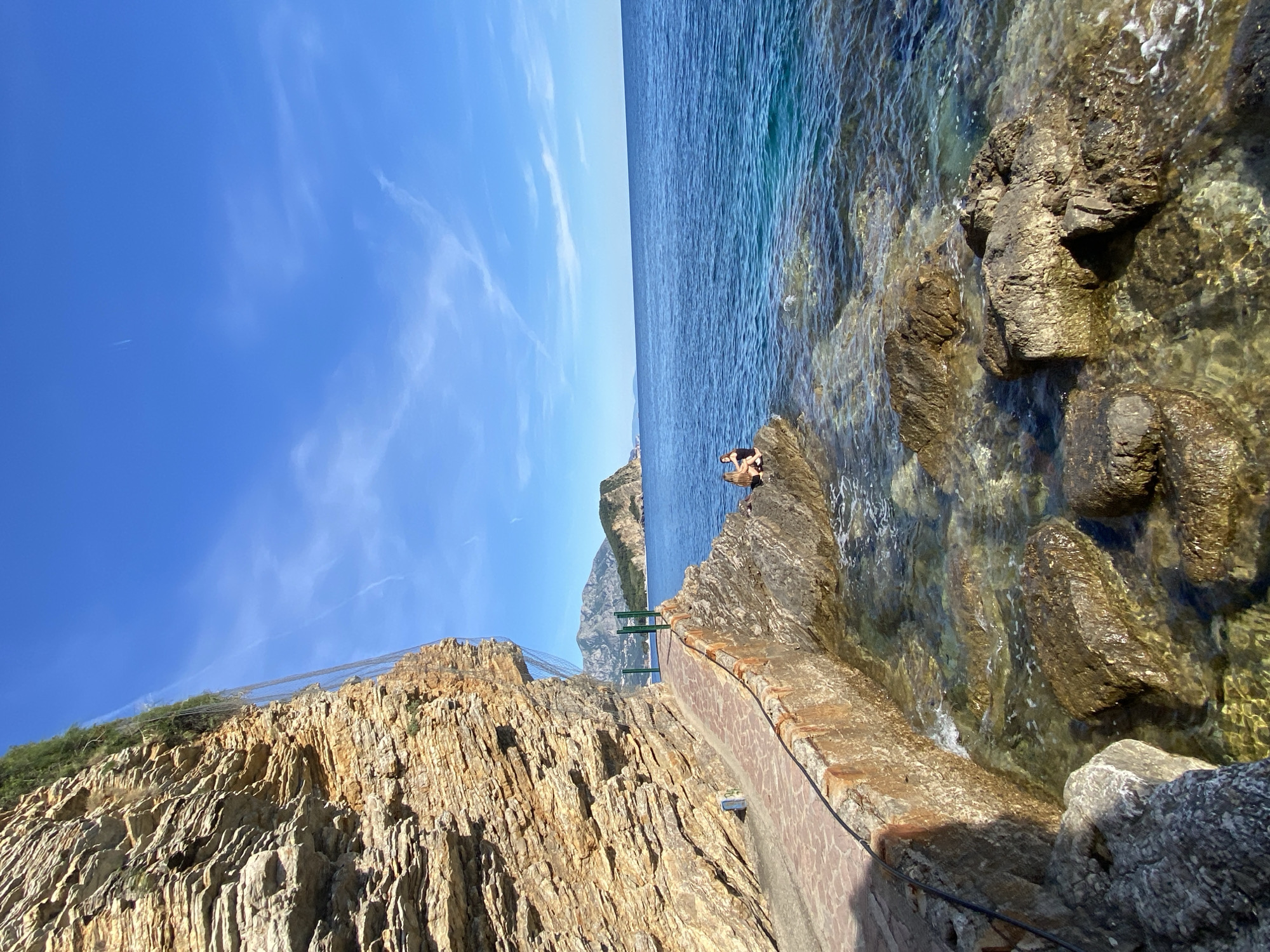 The image depicts a coastal scene in Budva, Montenegro. The sky is blue with wispy clouds above a calm, blue sea. Rocky cliffs form the shoreline, with a textured stone wall or path running along the edge. On the rocks at the water's edge, a person is sitting near the water. In the distance is a rocky mountain. A green metal structure rises from the path below. A safety net is visible on the side of the mountain. The water is clear, and one can see rocks beneath the surface.