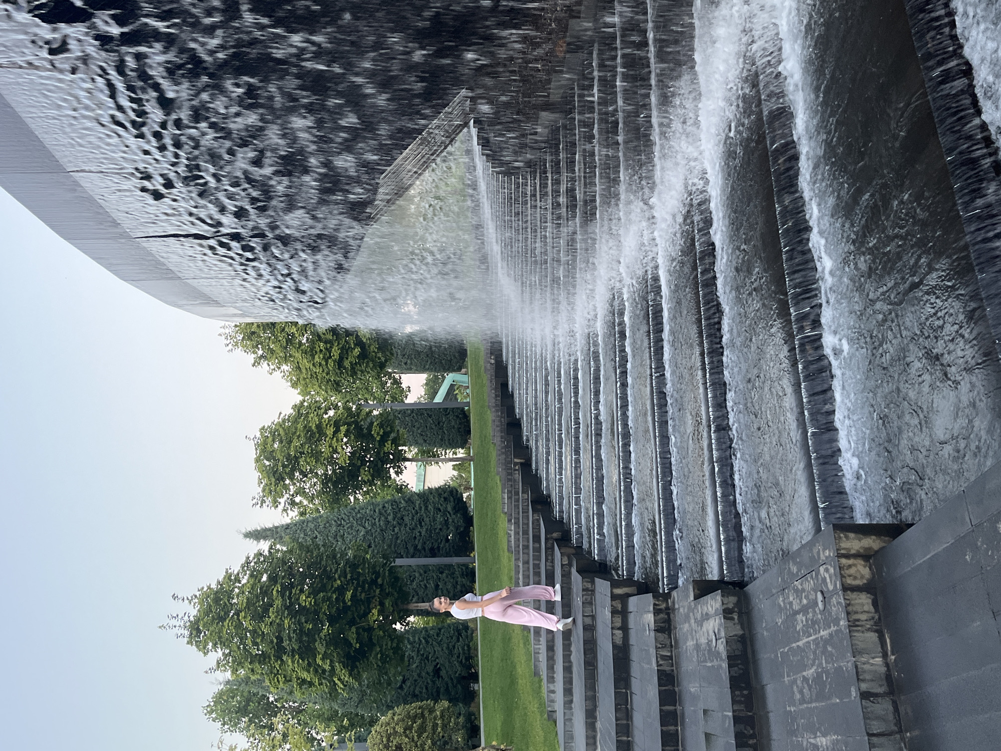 A young woman in pink pants and a white top stands on a stone staircase beside a cascading waterfall. The staircase is made of dark grey stone with a stepped design, and water flows down each level. The waterfall is a prominent feature, with water sheeting down a dark, curved surface. In the background, there are green trees and a light sky. The location is the "Solaris" fountain park in Krasnodar, Russia.