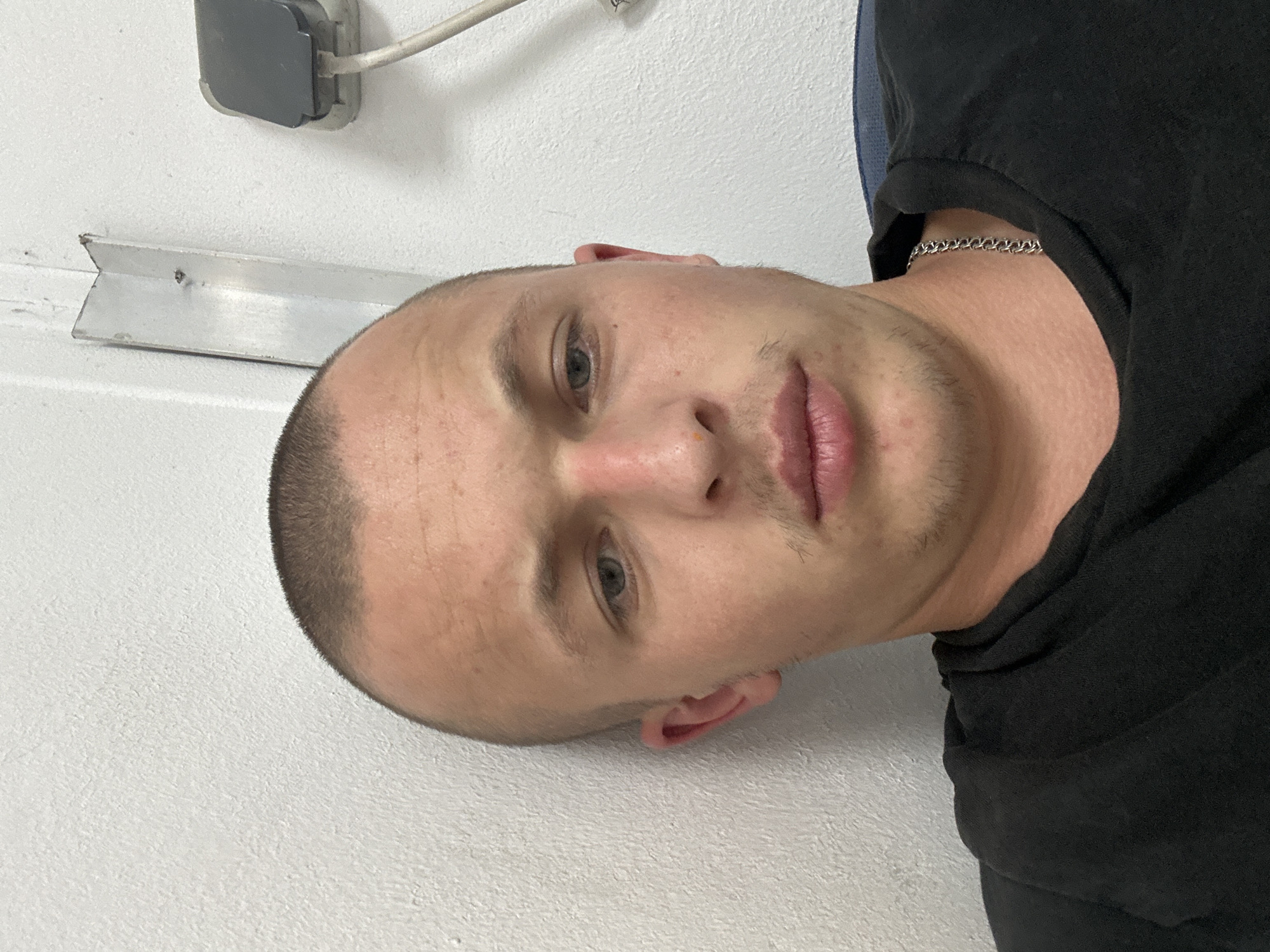 A male subject with a shaved head and light skin tone is shown in a close-up portrait. He is wearing a black t-shirt and a silver chain necklace. The background consists of a white wall with some industrial elements visible, including a metal corner bracket and an electrical outlet with a cord. The lighting suggests an indoor setting. The image does not provide any specific information about the location within the city of Mainz, Germany.