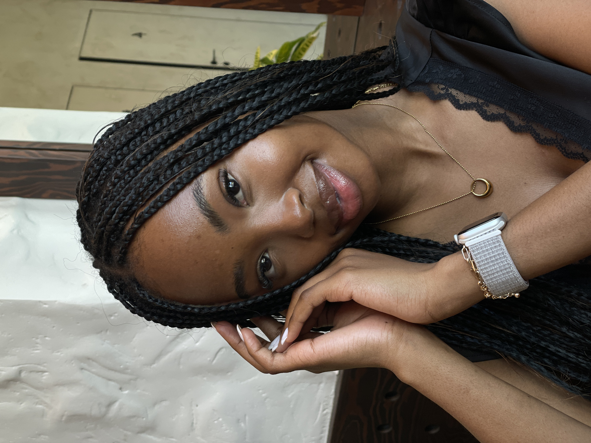 A close-up medium shot shows a young Black woman with braided hair, smiling at the camera. She is wearing a black top with lace trim, a gold necklace with a circular pendant, and a silver smartwatch with a light gray band. Her hands are positioned near her face, with one hand touching her braids and the other resting on her chin. The background is blurred, suggesting an indoor setting with light-colored textured walls and dark wood accents. The scene appears to be set in a restaurant or cafe.
