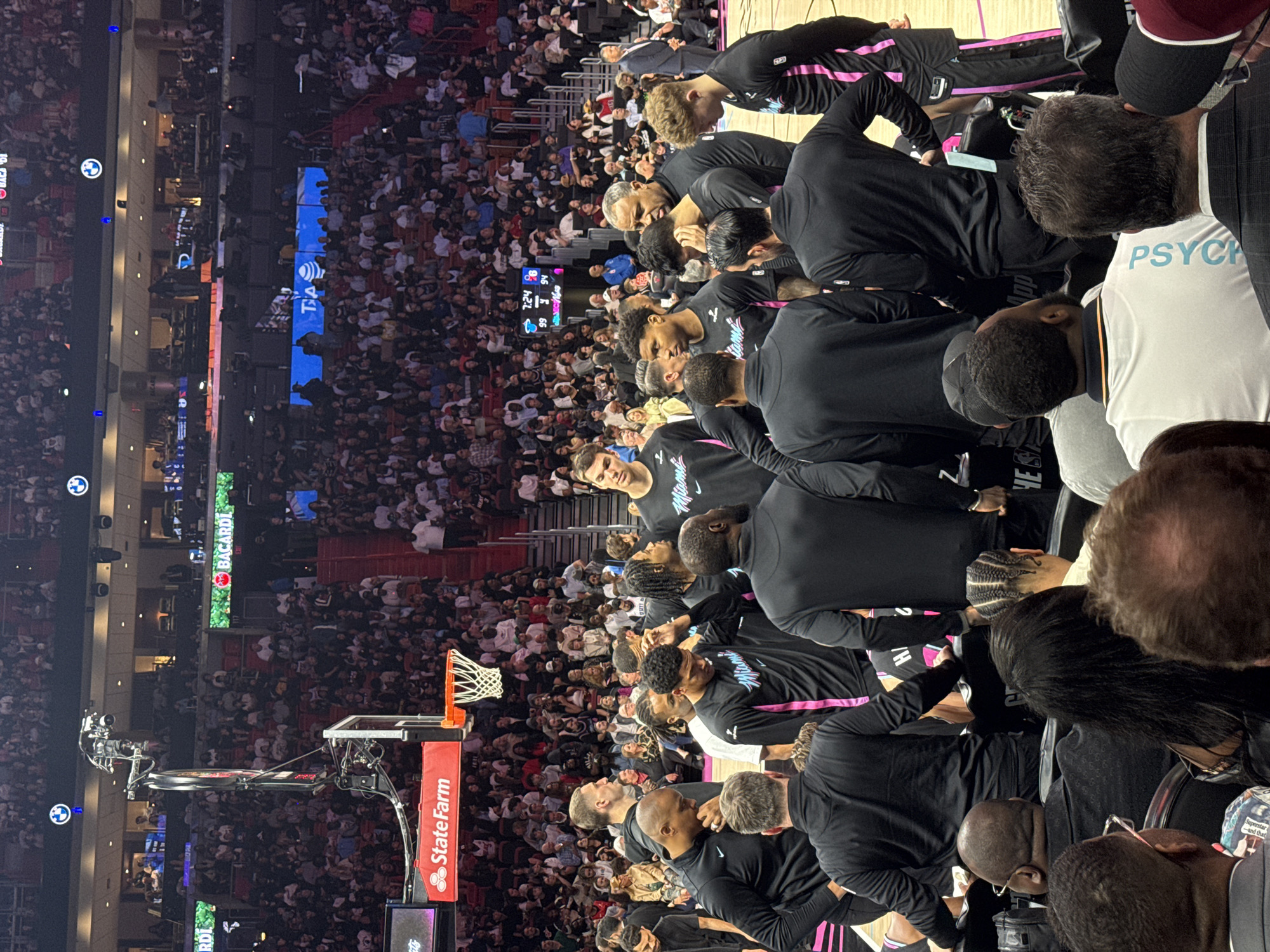 The image shows a crowded basketball game, likely the Miami Heat, given the team's distinctive black jerseys with pink accents, located in Miami, United States. The basketball court is visible with "State Farm" displayed on the side. Many of the spectators are seated in the stands. The coach's assistant staff also surrounds the team. One man is seen with a white shirt and the word "PSYCH".