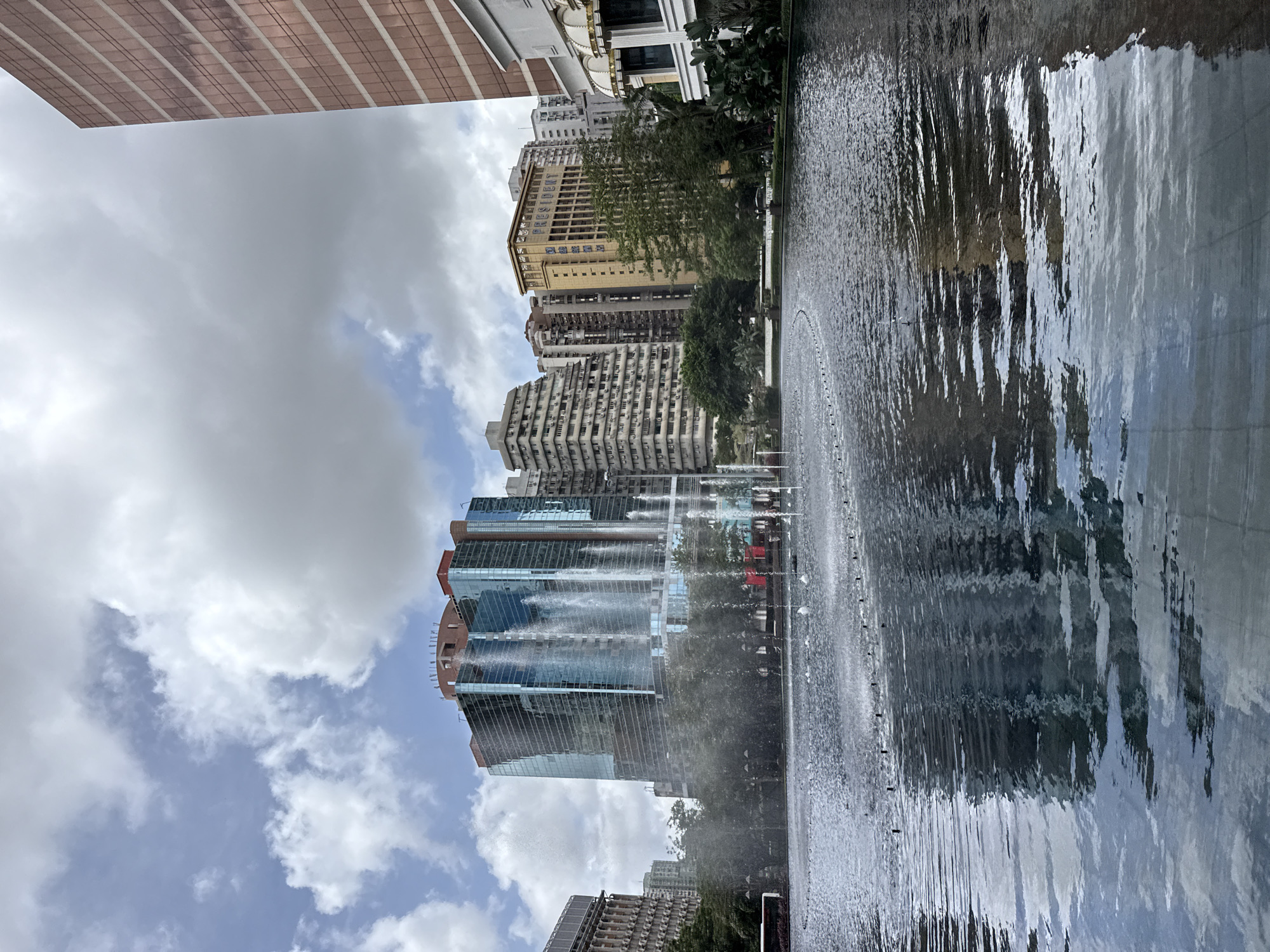 The image captures a wide view of the Wynn Macau resort's central performance lake, a prominent landmark in the city of Macau, China. The lake's surface is disturbed by numerous small water jets, creating a rippling effect. In the background, the iconic architecture of the resort is visible, including the main building with its distinctive golden hue and the glass facade of the Wynn Tower, where the famous "Tree of Prosperity" and "Dragon of Fortune" shows take place. The sky above is partly cloudy, with patches of blue sky peeking through. The scene is bustling with activity, as indicated by the presence of people walking around the lake and the general ambiance of a popular tourist destination. The architecture is a blend of modern and opulent styles, characteristic of Macau's integrated resorts, with a focus on grandeur and visual spectacle.