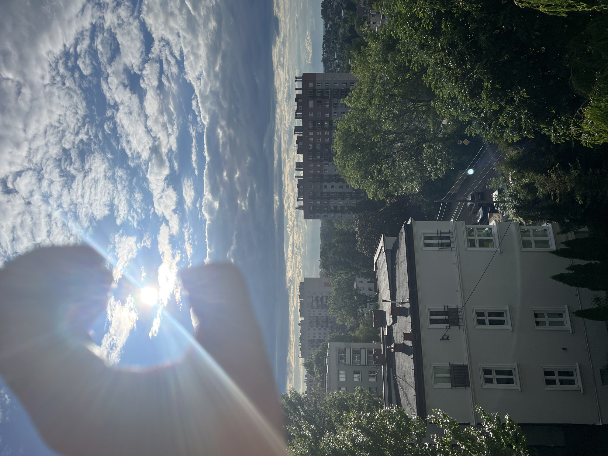 This is a high-angle, medium shot of a city skyline on a cloudy day. The sun is shining brightly through a gap in the clouds, creating a lens flare effect that dominates the top left portion of the frame. The lens flare is partially obscured by a blurred human hand, forming a heart shape with the thumb and index finger. Below the sky, several apartment buildings and trees are visible. The foreground features the roof of a white apartment building with chimneys. In the mid-ground, a larger, red and white apartment building stands out. The background consists of more residential buildings and a dense canopy of green trees, suggesting a suburban or urban environment. A street with a few parked cars is visible between the mid-ground buildings.