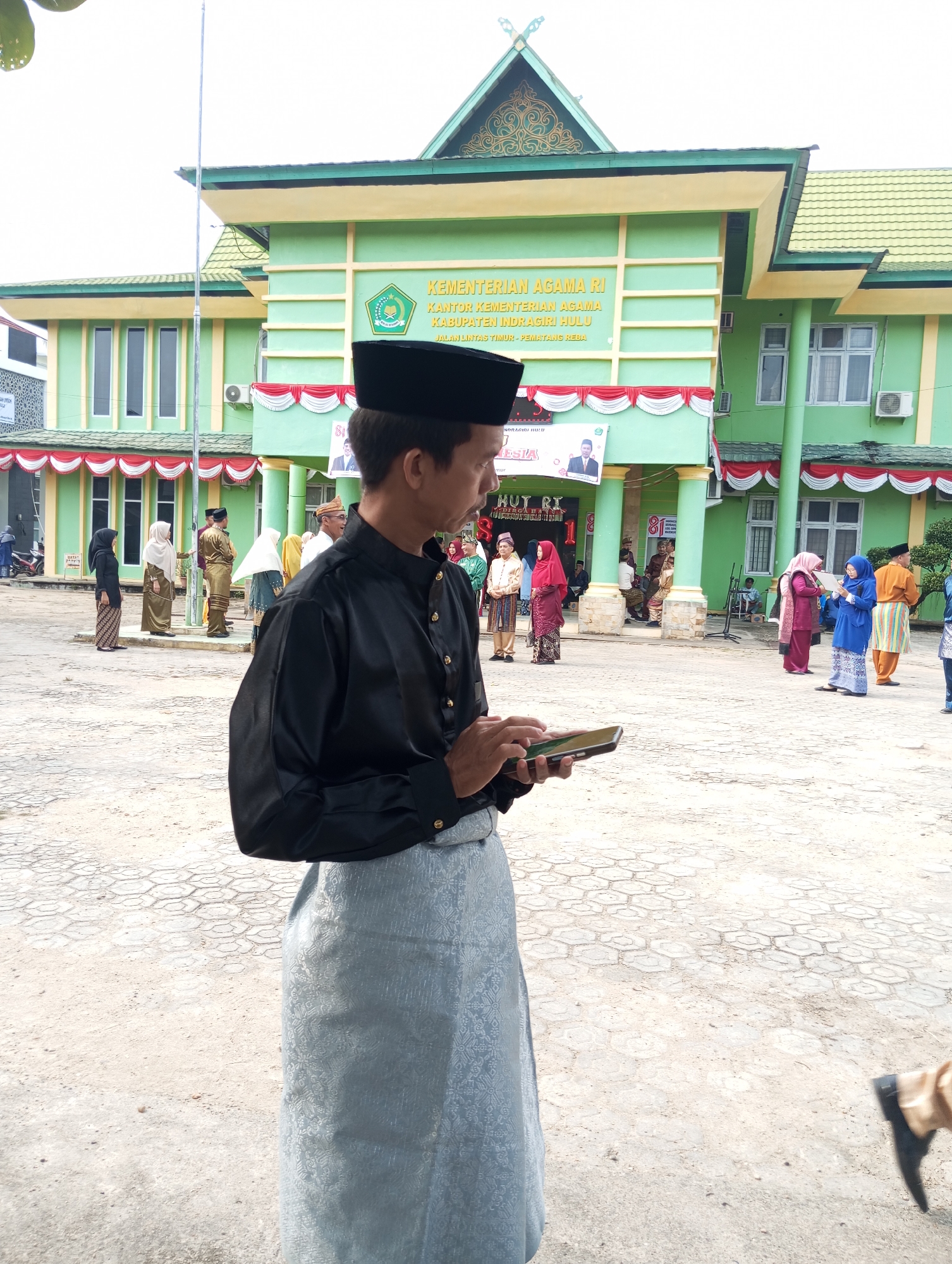 In front of a lime green building with yellow trim, a man wearing a black shirt, black peci hat, and a light blue patterned sarong holds a smartphone. The building has a prominent sign that reads "KEMENTERIAN AGAMA RI KANTOR KEMENTERIAN AGAMA KABUPATEN INDRAGIRI HULU JALAN LINTAS TIMUR - PEMATANG REBA." Several other individuals in traditional attire, including women wearing headscarves in various colors like red, blue, and beige, and men in traditional shirts and sarongs, are visible in the background. Some are standing and conversing, while others appear to be observing an event. The ground is paved with light-colored hexagonal tiles. A flagpole stands in the middle ground.