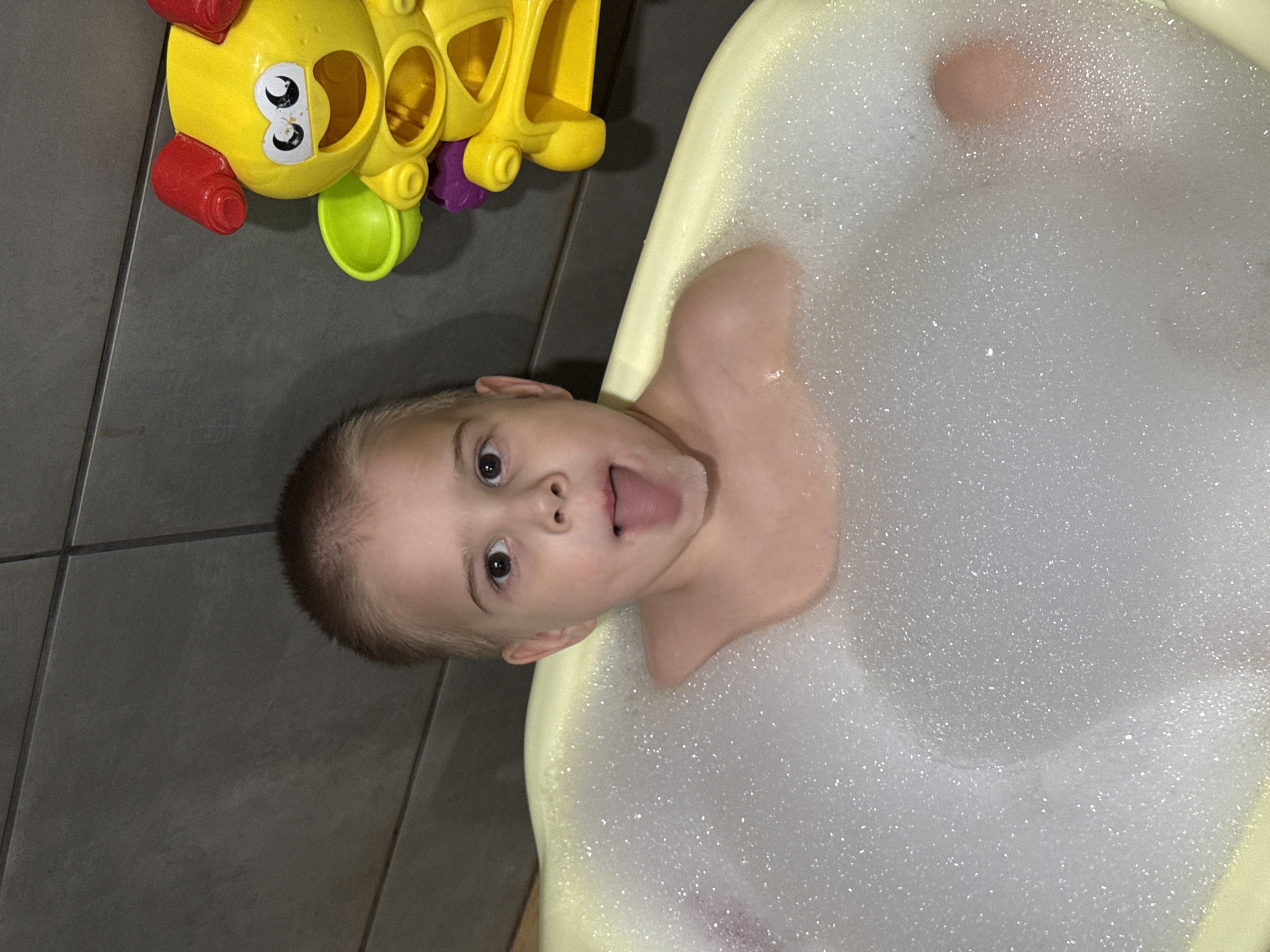 A child is in a bathtub filled with bubbles. The bathtub is light yellow. The child's head and shoulders are visible above the bubbles. The child has short, light brown hair and is looking at the camera with their mouth open. A yellow toy with colorful accents is on the floor next to the tub. The floor is tiled in gray. The scene is lit with artificial light. It is not possible to determine the exact location without additional information.