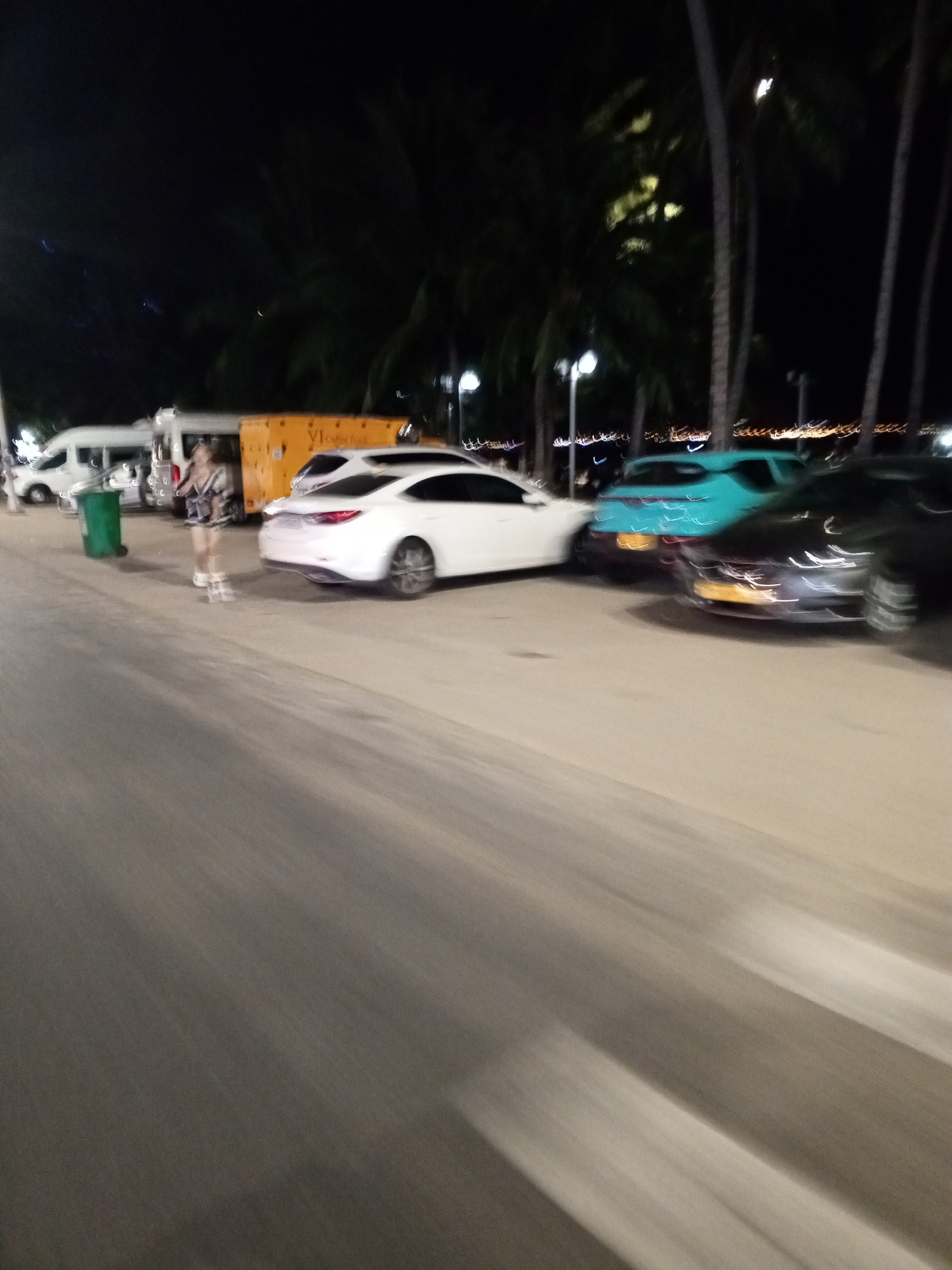 The media file depicts a nighttime scene on a street, likely in a tropical location, given the presence of palm trees. The overall image is blurred due to motion.
In the foreground, a person, appearing to be a woman, is standing. She is wearing a patterned outfit and white shoes. To her left is a green trash bin. Behind her, several vehicles are visible. A white van is parked on the left, followed by an orange food truck with "VI COFFEE TRUCK" written on it. Further to the right, a white sedan is parked, and beyond that, a teal-colored vehicle and a dark-colored car are also present. The background shows more palm trees and streetlights, suggesting an urban or tourist area. The lighting indicates it is nighttime. The blurred nature of the image implies movement, possibly from a moving vehicle or the photographer walking. The setting appears to be a street with parked vehicles and pedestrian activity. The overall mood is unclear due to the blur, but the presence of palm trees and what looks like a coastal setting might suggest a vacation or leisure destination.
