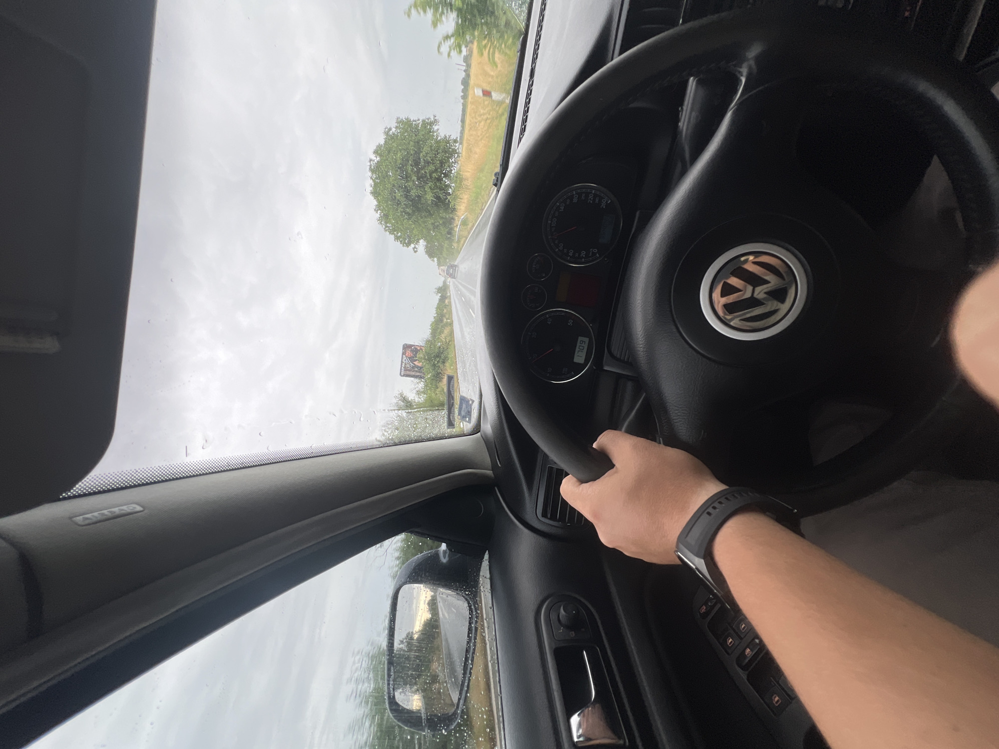 A first-person perspective shot from inside a car on a cloudy day shows the driver's hands on the steering wheel. The car's dashboard is visible, including the speedometer, which indicates a speed of approximately 110-120 km/h. The steering wheel has the Volkswagen logo in the center. Raindrops are visible on the windshield and side mirror, suggesting it is raining. The side mirror reflects the road ahead, which appears wet and is lined with trees and fields. In the distance, other vehicles can be seen on the road. A billboard advertisement is visible on the left side of the road. The location is identified as Cheshnegirovo, Bulgaria, based on the metadata.