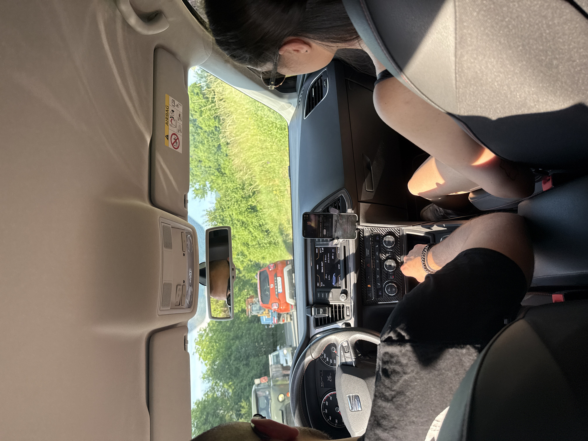 The image depicts the interior of a SEAT passenger vehicle, specifically a SEAT Leon (likely a 5F generation), with a clear view through the windshield onto a rural road in Novi Iskar, Bulgaria.
**People:**
Two individuals are present in the front seats. The driver, a male seen from behind, wears a black short-sleeved t-shirt. His right arm, adorned with two beaded bracelets, rests near the gear selector. His left hand is on the multi-function steering wheel, which features red stitching and a SEAT logo. The instrument cluster shows "10:25 AM" and a speed of 0, with a digital readout displaying "26". The front passenger, a female seen from behind, has dark hair and wears sunglasses on her head over a dark-colored top. Her legs are visible, illuminated by sunlight. The passenger seatbelt is fastened.
**Vehicle Interior:**
The car's interior features a dark dashboard with a carbon fiber-patterned trim around the infotainment system and climate controls. The infotainment screen displays "since start" data, including "0.1 km" driven, "0 km/h" average speed, and "0h 1m" driving time, indicating the vehicle is stationary or has just started. A smartphone, identified as an iPhone, is mounted vertically to the right of the infotainment screen, displaying an active interface. The rear-view mirror reflects the driver's forehead. Both sun visors are retracted; the passenger-side visor displays an "AIRBAG" warning label. The headliner is light beige/grey.
**Exterior Environment:**
The vehicle is on an asphalt road lined with dense green trees and tall grass on both sides under a clear blue sky, suggesting bright daytime conditions with strong sunlight. Traffic is visible ahead. Directly in front is an orange Dacia Duster SUV with a Bulgarian license plate partially visible. Behind the Duster, a yellow construction excavator/backhoe is present. To the left, a vintage-style green military-type truck, possibly a UAZ-452, is visible, with a white car behind it. The road appears to be a two-lane thoroughfare, possibly experiencing a traffic slowdown or queue.
