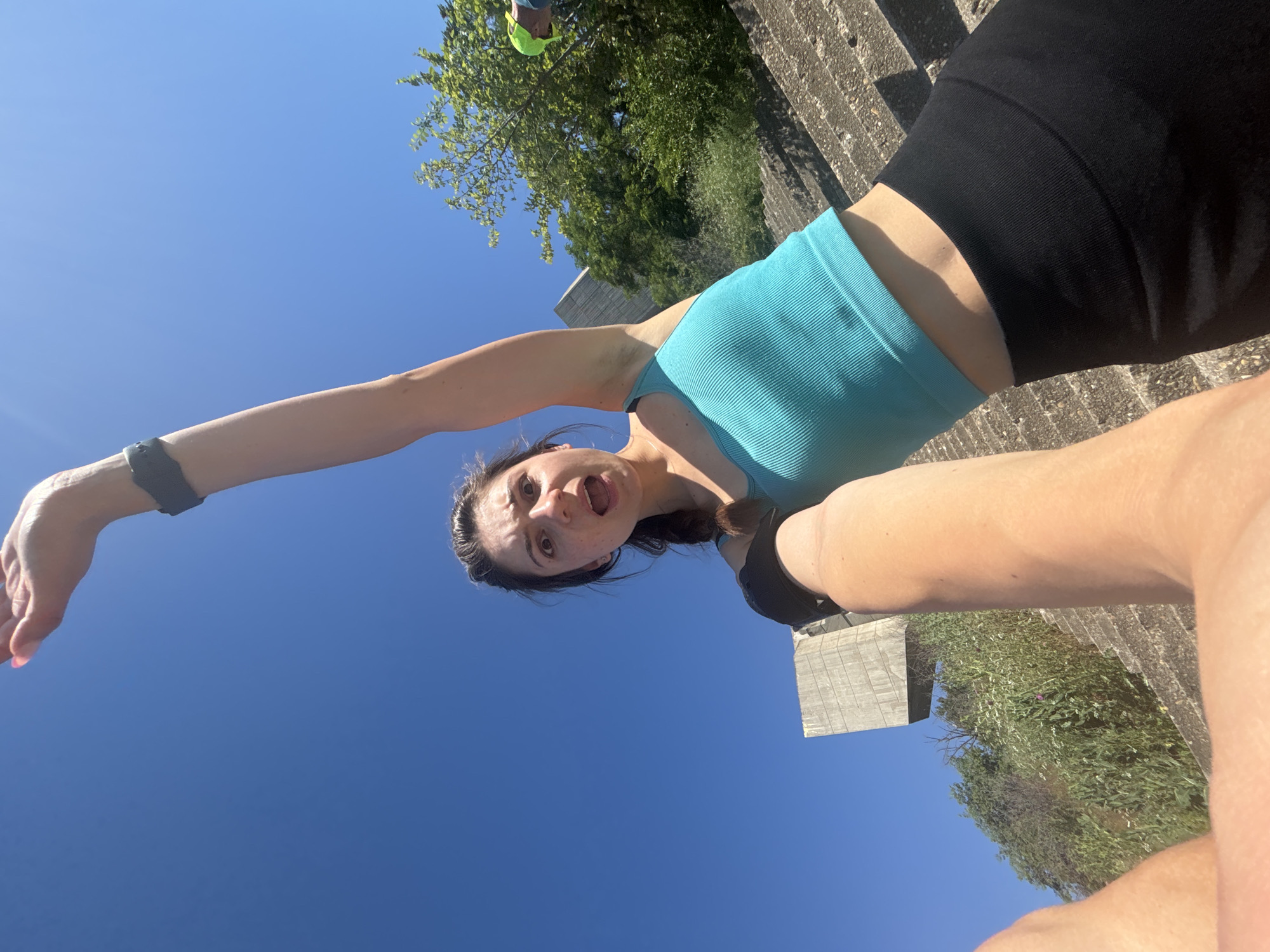 The image shows a young woman with brown hair in a braid, wearing a turquoise tank top and black leggings. She is taking a selfie with an outstretched arm, looking directly at the camera with an open mouth and surprised expression. Her right arm is raised upwards, with a black fitness tracker on her wrist. The background features a bright blue sky, lush green trees, and a large, concrete structure that appears to be part of a monument. The woman is positioned on a set of concrete stairs, and there are hints of vegetation and other people in the distance. The concrete structure in the background is the Buzludzha Monument, a significant landmark in Bulgaria.