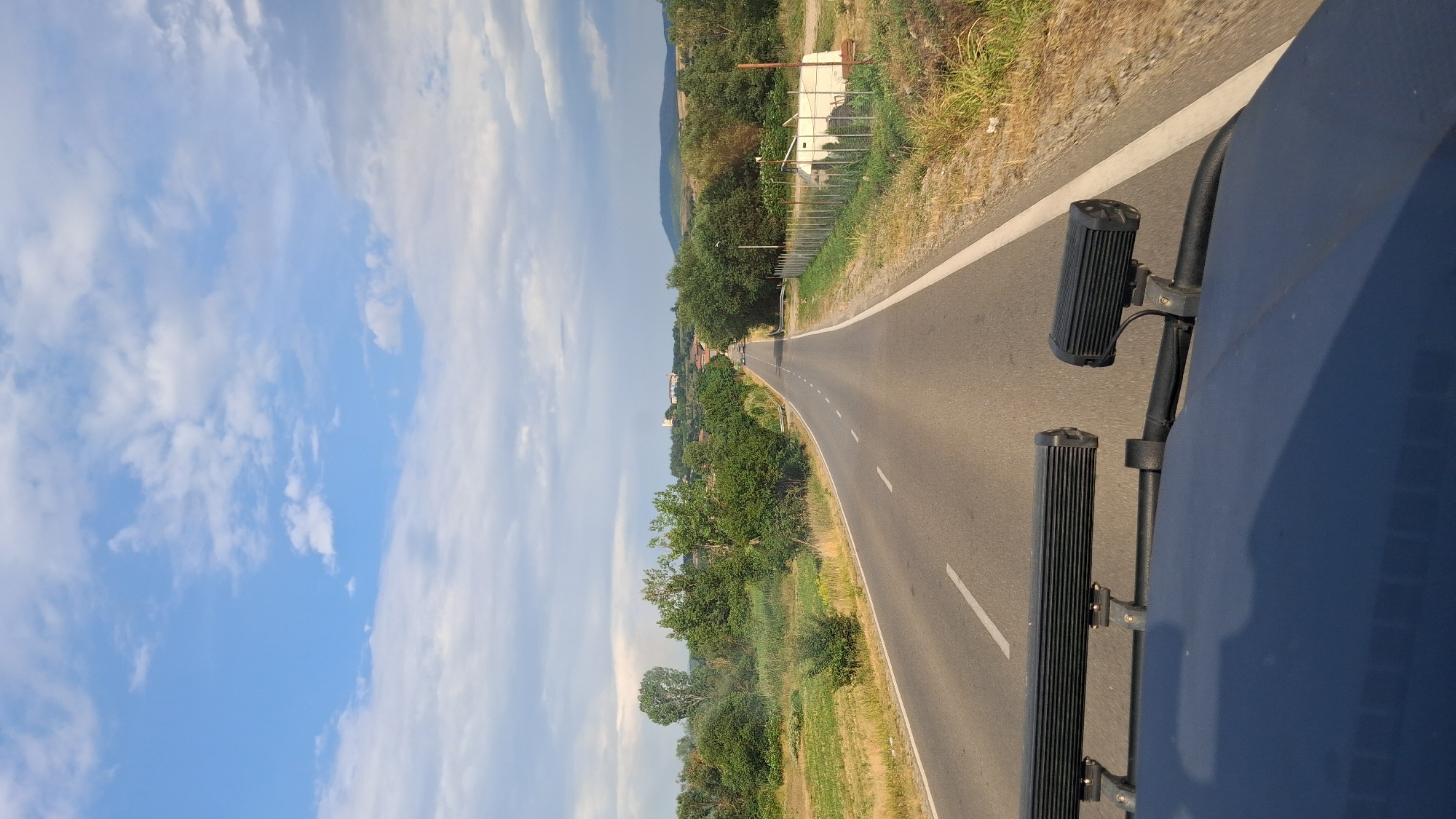 The image depicts a view from the front of a vehicle driving on a paved road. The road curves gently to the left and extends into the distance, where a town or village is visible on a hill. The landscape on either side of the road is rural, with fields and trees. The sky is partly cloudy, with patches of blue and white. Two auxiliary lights are mounted on the roof rack of the vehicle in the foreground. There are no people visible in the image. The scene is consistent with a drive through the countryside near Hadzhidimovo, Bulgaria.