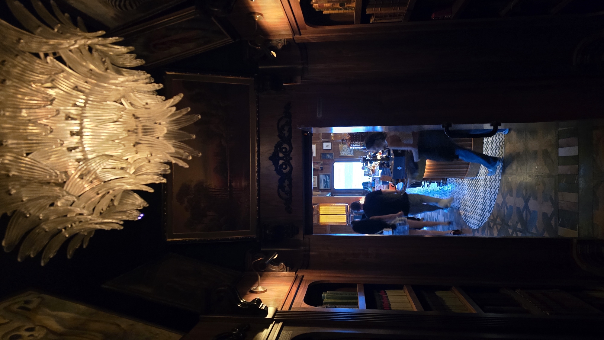 The image depicts the interior of a dimly lit establishment, possibly a bar or restaurant, with ornate wooden paneling and a large, elaborate chandelier hanging from the ceiling. In the foreground, to the left, is a bookshelf filled with books. To the right, the view opens into a wider area where several individuals are present. Two people are walking away from the camera towards a bar area, one a woman with dark hair, the other a man wearing a dark t-shirt. Another person, a woman with blonde hair, is seen in the mid-ground to the right, facing away from the camera and towards the bar. The floor has a patterned tile design. The overall ambiance is one of old-world charm and luxury. Based on contextual information not present in the image itself, this scene is located in San Diego, United States of America.