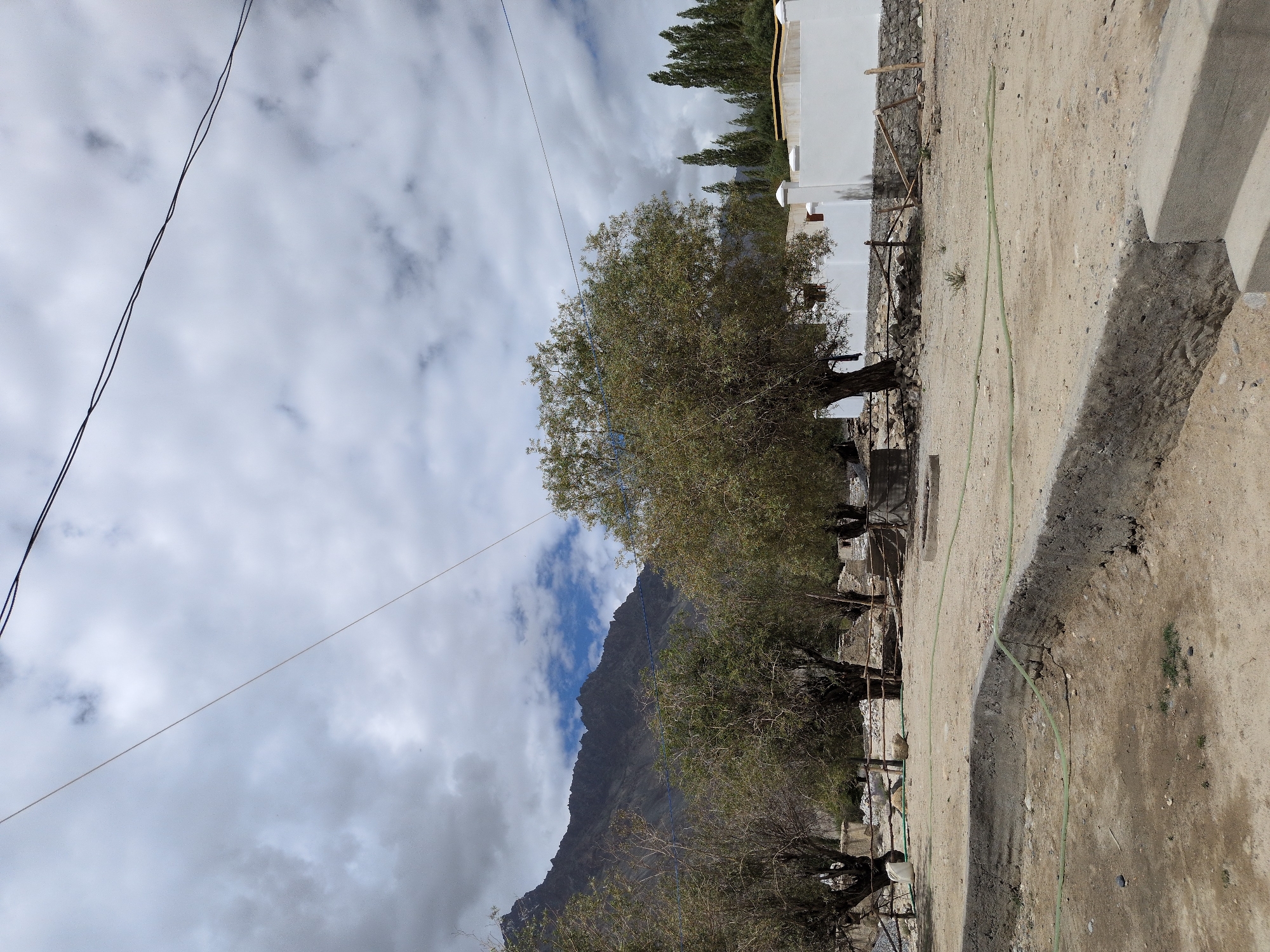 This is a full, eye-level, outdoor shot of an arid landscape in Panamik, India, on a cloudy day. In the foreground is a dry, dirt-floored area with a concrete structure. A green hose snakes across the ground. In the middle ground are several trees with sparse foliage, and a section of white wall with a fence. In the background are barren, mountainous peaks under a dramatic, cloudy sky. Power lines stretch across the top of the frame.
