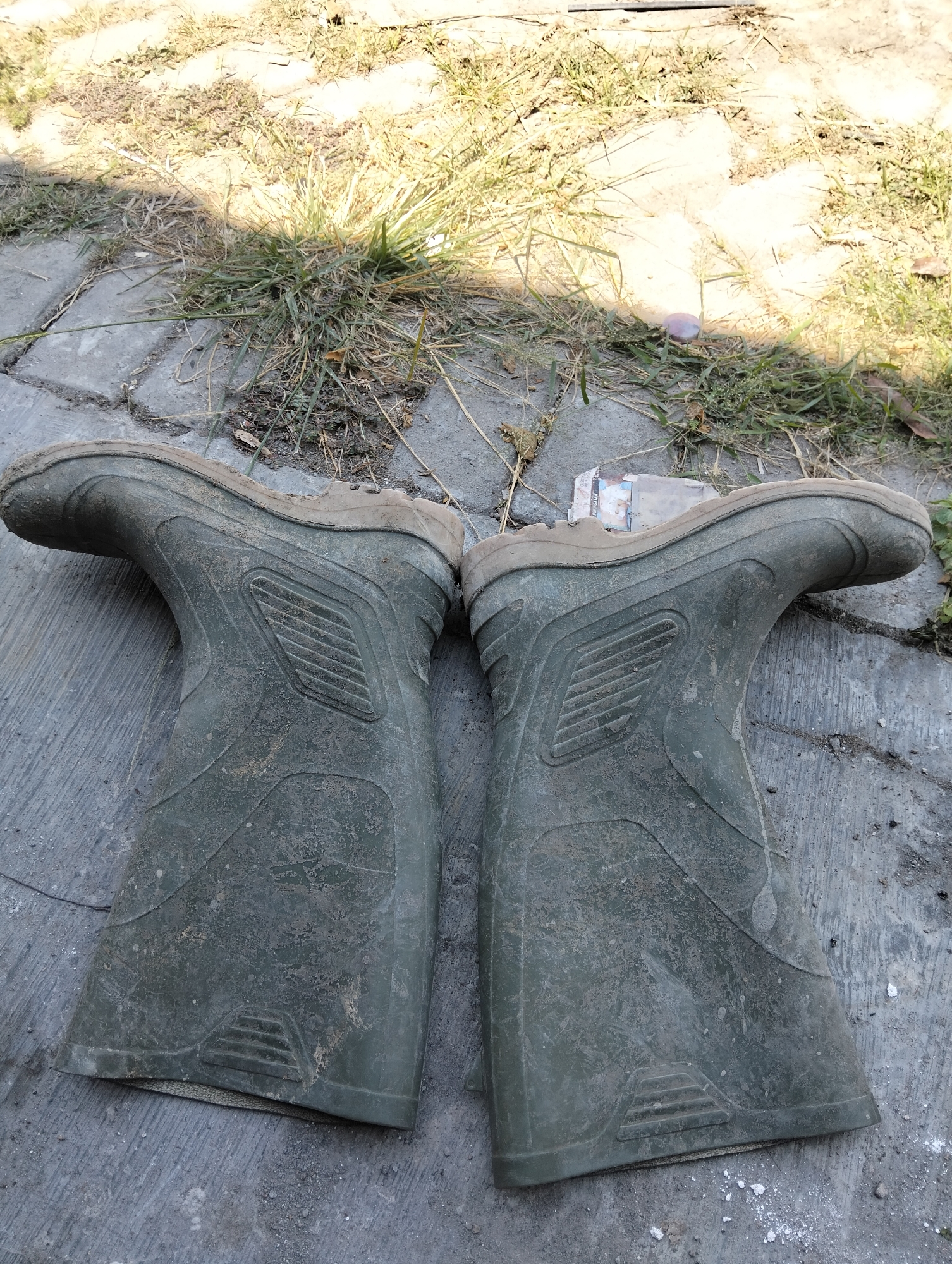 A pair of dirty, green rubber boots are laid out on a cobblestone surface. The boots are facing away from the camera and are positioned side-by-side. They appear to be gardening boots or work boots, judging by their sturdy construction and the mud caked on their surfaces. The cobblestones are weathered and uneven, with patches of dry grass and dirt growing in the gaps between them. A shadow from an unseen object falls across the left boot and the surrounding area, suggesting it is daytime. The background is blurred, but it appears to be an outdoor environment with more grass and sunlight. The scene is devoid of human presence, and no specific actions are being depicted. The overall impression is one of a functional, utilitarian space, possibly a backyard or a work area. There is no information in the image to indicate this location is in Semarang, Indonesia.
