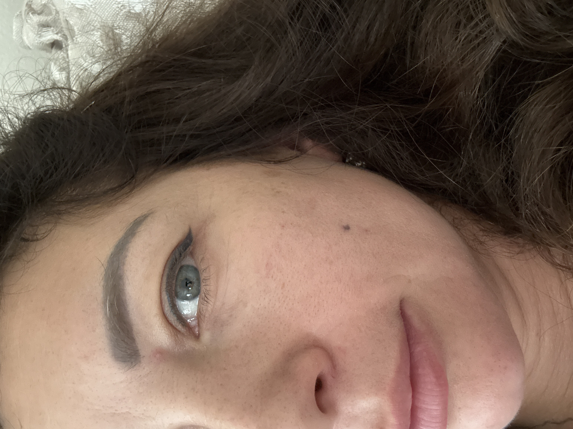 The image is a close-up, horizontally oriented, personal photograph focused on a portion of a person's face. The subject appears to be a young woman with brown, wavy hair that frames her face and extends out of frame. Her left eye, with a light blue iris and visible eyelashes, is a prominent feature, accented by dark eyeliner forming a subtle wing. Her eyebrow is groomed and shaped. The skin is fair with some visible pores and freckles. There is a small, dark beauty mark above her cheekbone, near the temple. A portion of her nose and lips, which are painted with a pink lipstick, are also visible. The background is out of focus, showing hints of a textured, possibly fabric surface, in a light color, and more of the subject's hair. The lighting suggests an indoor setting. The image appears to be a selfie, given the close-up perspective. No specific activity is taking place; it is a posed or candid portrait. No text is visible in the image.