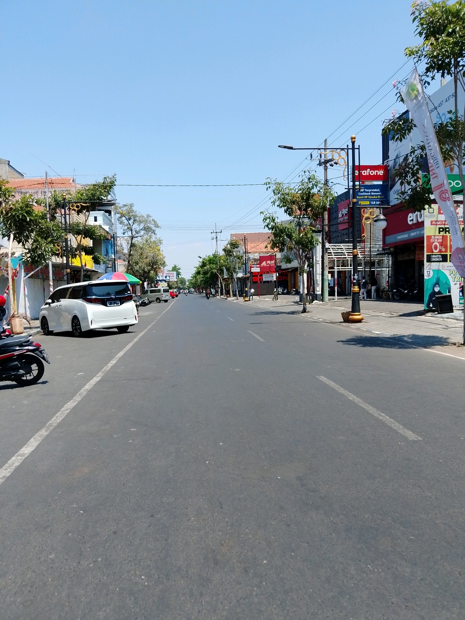 A street scene in Pasuruan, Indonesia, features a wide, paved road with a white line marking the lane. On the right side of the road, several businesses line the sidewalk, including a Vodafone store and a shop with a large red sign. A white minivan is parked on the left side of the road. Trees and utility poles are visible along the street. The sky is clear and blue.