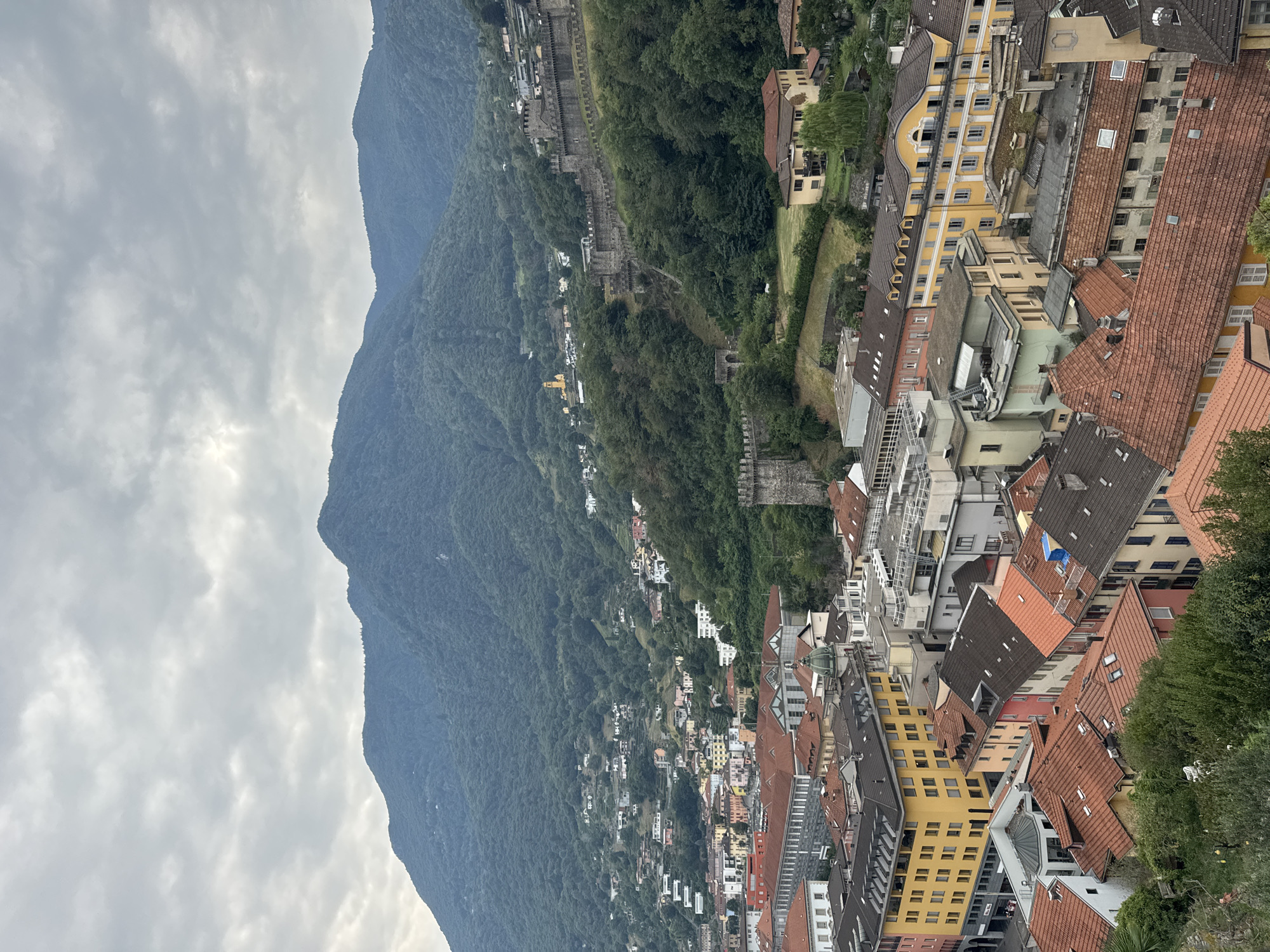 The image shows a panoramic view of Bellinzona, Switzerland, focusing on its historic castles and the surrounding landscape. In the foreground, the terracotta roofs of residential buildings dominate the lower half of the frame, with various architectural styles and colors. A prominent bright yellow building with arched windows stands out among the rooftops.
The middle ground reveals more of the urban sprawl, with buildings clinging to the hillside. A significant feature is the ancient stone wall of Castel Grande, a UNESCO World Heritage site, snaking its way up the verdant slope. Several defensive towers and fortifications are visible along the wall. Further up the mountain, more scattered houses and the yellow spire of a church can be seen.
The background is dominated by a large, densely forested mountain that rises majestically. The sky is overcast with heavy, grey clouds, creating a diffused light that casts a soft, somewhat muted tone over the entire scene. The overall impression is one of a historic European town nestled in a dramatic natural setting. No people are clearly discernible in the image.