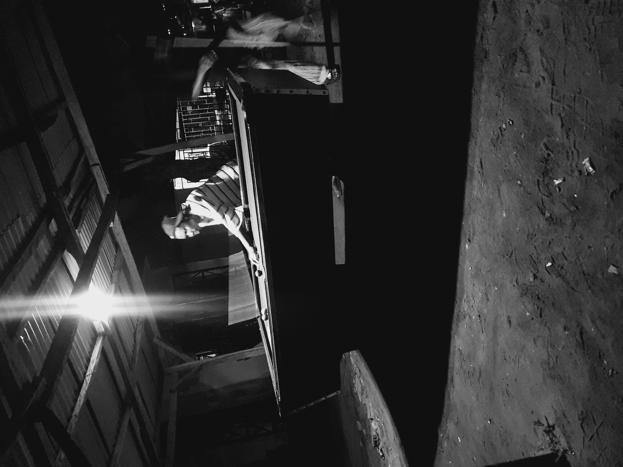 An indoor scene, heavily contrasted in black and white. A man wearing a striped shirt leans out a window. Another individual, apparently in motion, stands behind him on the outside of the building. The building appears to be made of wood. A light source, either natural or artificial, shines through an opening, causing bright streaks. A concrete wall or walkway is on the right. The location is unknown, but speculated to be Lokoja, Nigeria.