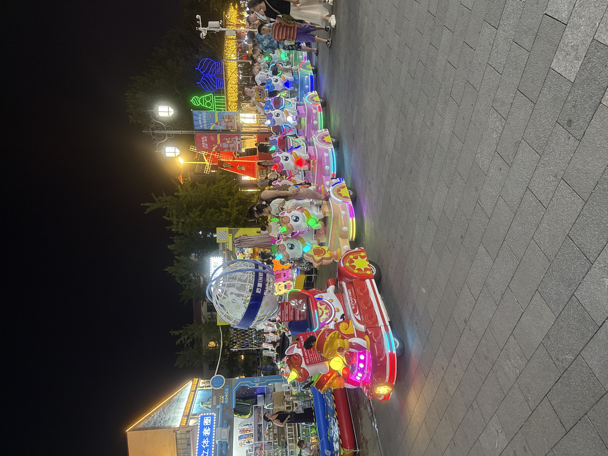 This image depicts an amusement ride at night in Qinhuangdao, China, likely in an outdoor pedestrian zone or entertainment area. The primary focus is a colorful, illuminated train-like amusement ride with individual cars shaped like unicorns and other cartoon animals. The ride is moving, with several children and adults seated in the cars. The ride is predominantly pink, white, and red, with bright LED lights. In the background, there are other amusement attractions, including a windmill-themed structure and illuminated decorative arches with onion domes. Several people are visible in the background, some appearing to be patrons of the amusement area, while others may be staff. The ground is paved with rectangular stone tiles. To the left of the main ride, there is a booth or stall with merchandise displayed, and a woman is visible near a blue pool. The overall atmosphere is festive and lively, characteristic of a nighttime entertainment venue. The image captures a typical scene from a popular tourist or recreational spot in Qinhuangdao during the evening.