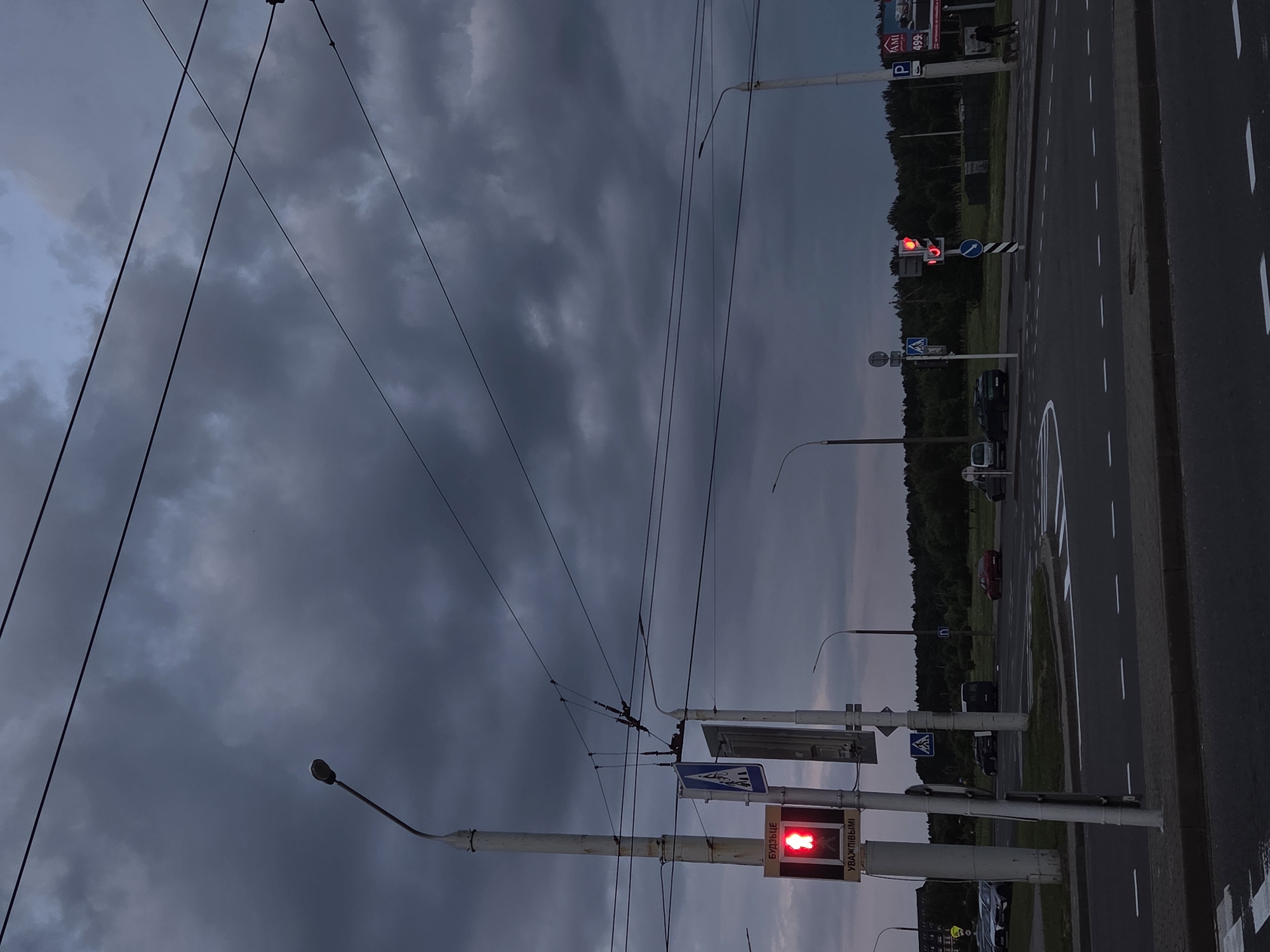 The image depicts a cloudy sky over a street intersection in Minsk, Belarus. A red pedestrian signal is illuminated on the left side of the frame, indicating "Do Not Walk". Overhead power lines crisscross the sky, supported by tall lampposts. In the background, several cars are visible, along with a line of trees. A traffic light on the right side of the intersection shows a red light for vehicles. The overall scene suggests dusk or an overcast day.
