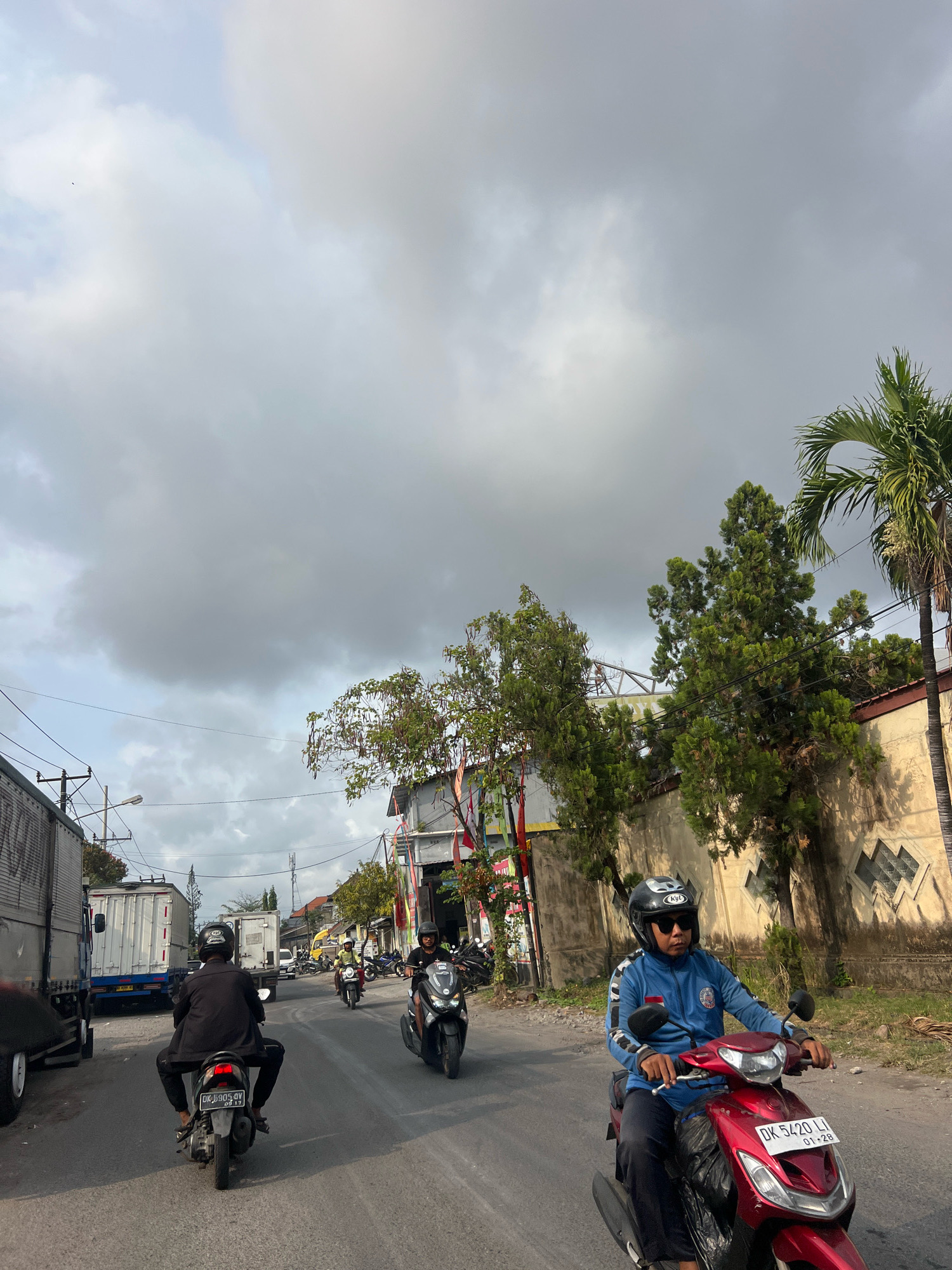 The image depicts a street scene in Denpasar, Indonesia, likely during daylight hours with overcast skies. The foreground shows two motorcycles traveling on a paved road. The motorcycle on the right is red, ridden by a person wearing a blue jacket, black helmet, and sunglasses. The license plate is visible and reads "DK 5420 LJ". The motorcycle on the left is black and being ridden by a person wearing a black jacket and helmet, with the license plate showing "DK 5905 QV".
In the middle ground, other motorcycles and a yellow car are visible, suggesting moderate traffic. Several trucks are also present, one white truck with a blue stripe on the left side of the road, and another white truck further down the road. The roadside features buildings with light-colored walls, some with decorative patterns. Several trees, including palm trees and other lush green foliage, are visible along the street, some reaching over the buildings. Power lines and street lights are also part of the infrastructure. The sky is heavily clouded, casting a diffused light on the scene. Some flags or banners are hung on poles along the street, adding a touch of local color.