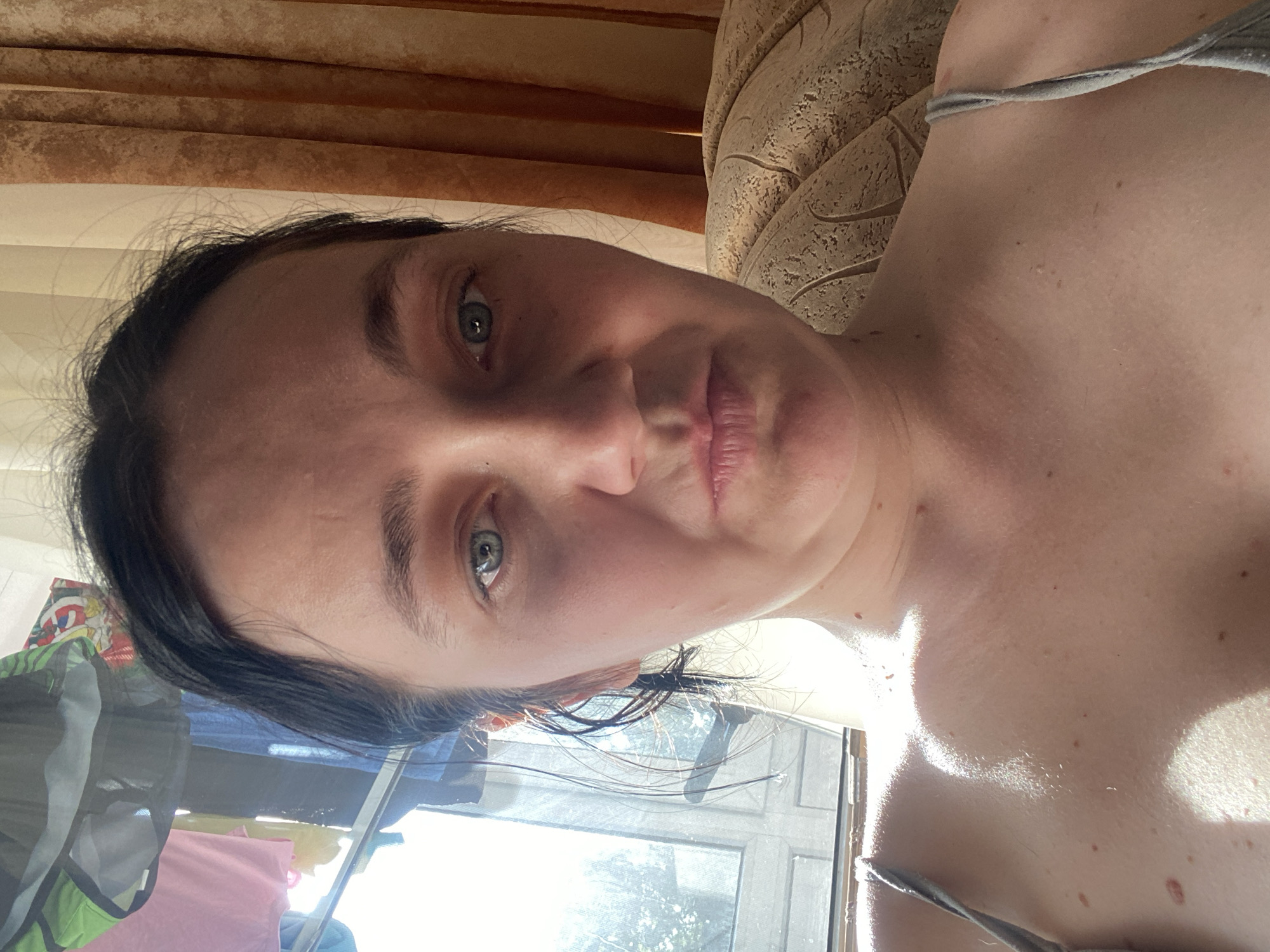 A medium shot, waist-up, shows a woman with dark hair, fair skin, and blue-green eyes. She is looking directly at the camera with a neutral expression. She is wearing a grey tank top, with straps visible over her shoulders. Her hair is wet and pulled back from her face, with some strands falling forward. The background is out of focus and shows a window with sunlight streaming through, casting shadows on her skin. There are curtains to the right of the frame. The lighting is natural and strong, creating harsh shadows. The overall impression is of a casual, indoor setting. The location is Unknown, Unknown.