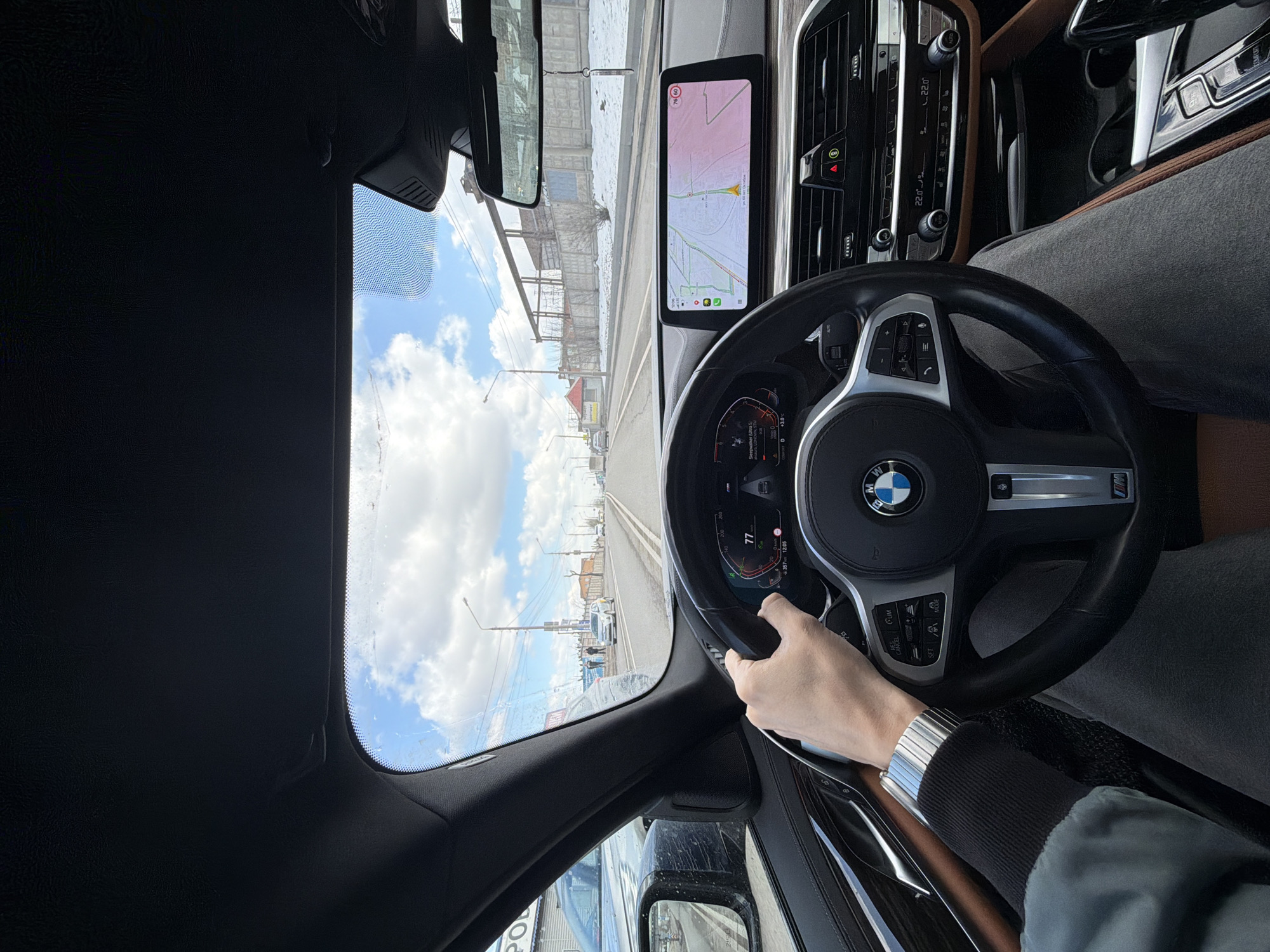 The image captures a driver's perspective from inside a BMW vehicle, looking forward through the windshield. The driver's right hand, adorned with a metallic wristwatch, is gripping the steering wheel, which features the BMW logo and an "M" badge. Visible steering wheel controls include "RES/CNCL," "SET," "LIM," and "MODE."
The car's dashboard displays a digital instrument cluster showing a speed of "77" (likely km/h), an outside temperature of "-3.0°C," and the time "12:05." To the right, a central infotainment screen shows a navigation map with Cyrillic text, indicating a location in Russia. Below the screen, the climate control panel is set to "22.0°C" with "AUTO" engaged. The interior features dark and brown trim.
Outside, the scene depicts a road under a partly cloudy, bright sky during daytime. The road has clear white markings, and streetlights line the side. Buildings, some appearing industrial, are visible in the distance, along with bare trees and bushes, suggesting a late winter or early spring season in Krasnoyarsk, Russia, as per the context. A building by the road has the Cyrillic text "СТО" displayed. A white car and other vehicles are present further down the road. The windshield shows some spots or grime.