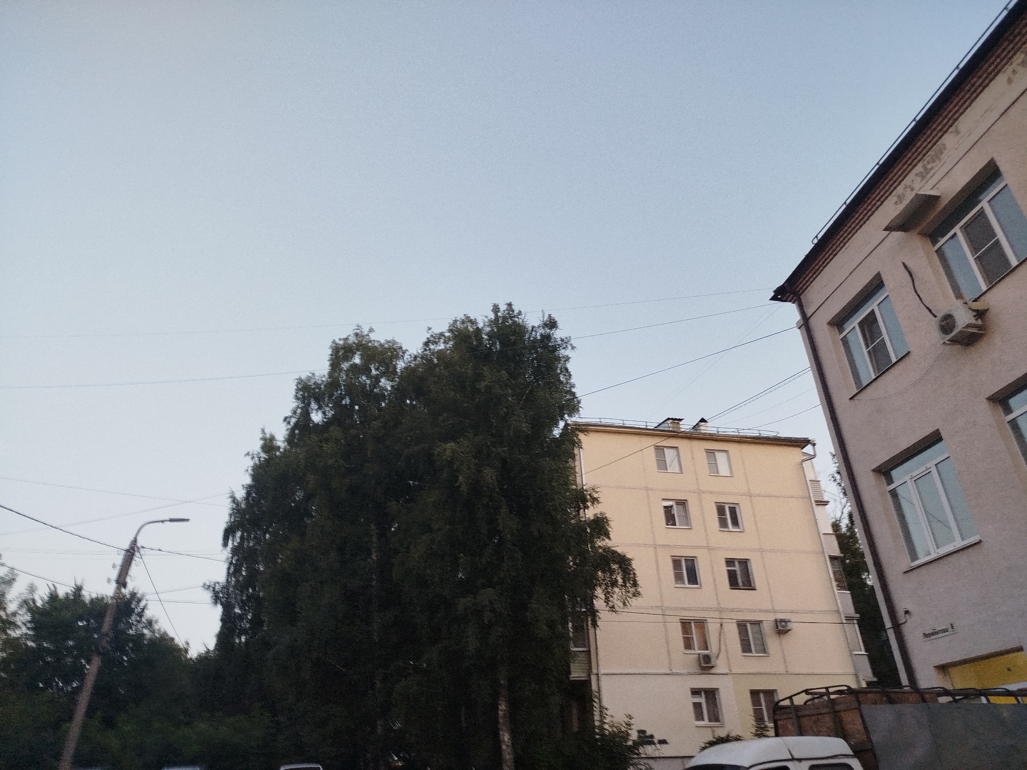 The image depicts a street view in Ryazan, Russia, featuring a light beige five-story apartment building. The building has several windows, and at least one visible air conditioning unit. To the left of this building, another, taller apartment building of a similar color is partially visible. In the foreground, a large, dark green tree with dense foliage is prominent. A street lamp with a curved arm and a light fixture is visible in the middle ground, along with electrical wires strung across the scene. Further to the right, a portion of another building, lighter in color, is seen with windows and what appears to be a sign on its facade. In the bottom right corner, part of a truck with a wooden cargo bed and a white vehicle are visible. The sky is a pale, clear blue, suggesting daytime.