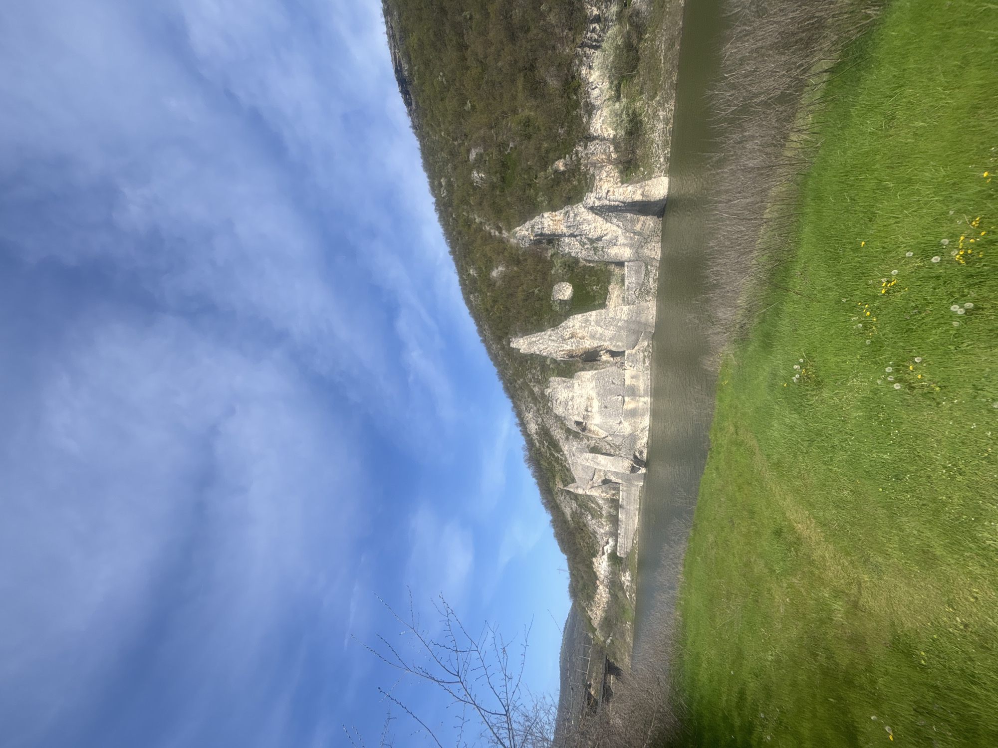 The image displays a scenic natural landscape, likely a river valley or gorge in Asparuhovo, Bulgaria, captured during the day.
A clear blue sky with scattered, wispy white clouds occupies the upper portion of the frame. Below the sky, a prominent and extensive hillside or mountain stretches horizontally across the mid-ground. This hillside is characterized by steep, light-colored rock formations, cliffs, and patches of vibrant green vegetation. At the foot of the hillside, a calm, dark greenish-brown body of water, consistent with a river or reservoir, extends horizontally.
Adjacent to the water on the right, a wide expanse of lush green grass is visible, adorned with small yellow and white wildflowers, suggesting a spring or early summer setting. Man-made structures, appearing as light-colored concrete or stone retaining walls, are integrated into the base of the rocky hillside near the water, possibly indicating a dam, bridge supports, or historical fortifications. In the bottom left foreground, the bare branches of a tree or bush are visible, further hinting at early spring.
The scene is brightly lit, implying clear weather conditions. There are no people, visible activities, or discernible text in the image, conveying a sense of serene natural beauty and tranquility.
