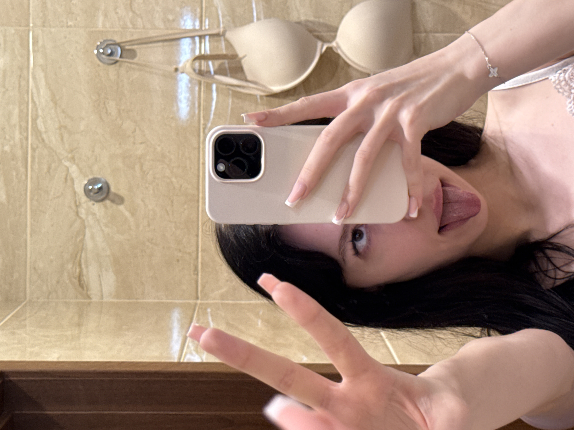 In a bathroom with beige tiled walls, a young woman with long dark hair takes a mirror selfie. She is holding a smartphone in her right hand, obscuring part of her face. Her left hand is raised, with two fingers extended in a peace sign. She is sticking her tongue out. A beige bra is hanging on hooks on the wall behind her. A delicate bracelet with a small star charm is visible on her wrist.