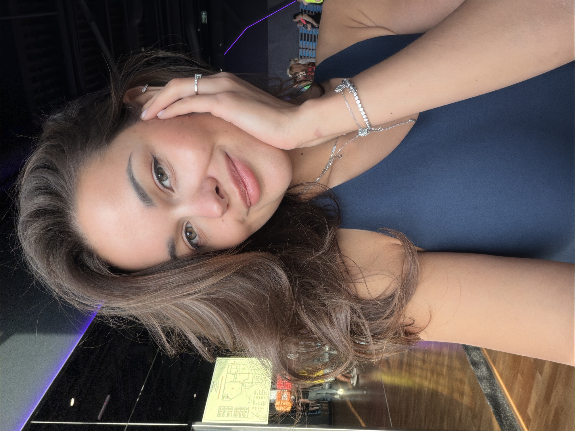 A portrait-style selfie features a young woman with medium-brown, wavy hair looking directly at the camera with a gentle, natural smile. Her head is slightly tilted, and her left hand is raised to lightly touch her temple and cheek. She has dark eyes and full lips. On her left ring finger, she wears a silver-colored ring with a prominent clear stone. Her right wrist is adorned with a sparkling silver-colored bracelet featuring numerous small clear stones, and a more delicate silver chain bracelet, possibly connected to a necklace with a rectangular pendant resting on her chest. She is wearing a dark blue sleeveless top.
The scene is set indoors, likely within a modern public facility such as a gym, sports complex, or entertainment center in Krasnoyarsk, Russia. The background is dark with ambient artificial lighting, including subtle purple-blue light accents on the left. Blurred figures of other people are visible further back, some appearing to be engaged in activities, suggesting a lively environment. A yellow-green map or floor plan diagram is visible on a reflective surface in the lower left background, confirming a public building setting. The floor in the foreground is dark and reflective, mirroring distorted shapes of the surroundings. The image conveys a casual, candid moment under artificial lighting conditions. No specific text is clearly legible, only indistinct details on the background map.